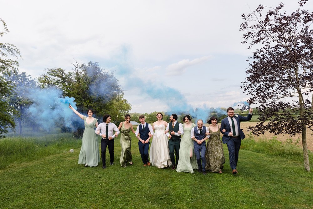 Bridal party walking through the field at Huntsmill Farm with blue smoke bombs, Buckinghamshire countryside beyond.