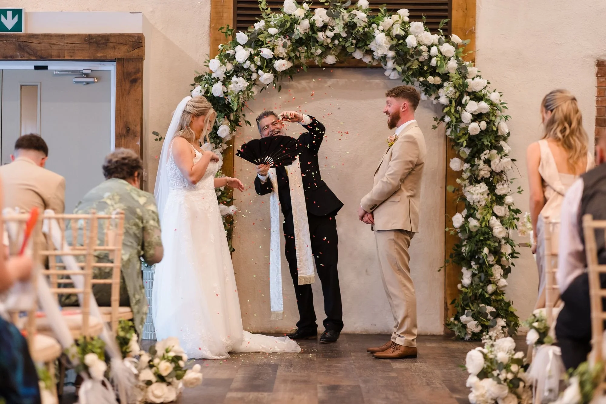Celebrant turning a paper rose into a burst of confetti between Abbie and Sean under a white rose arch in the barn at Bennetts Willow Barn