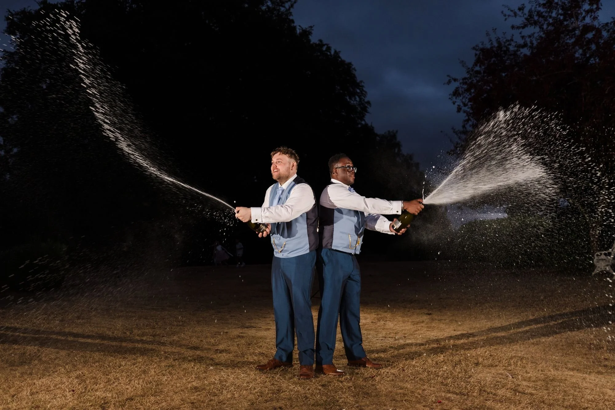 Liam and Shaun standing back to back on the lawn at night, each spraying a bottle of champagne into the dark sky