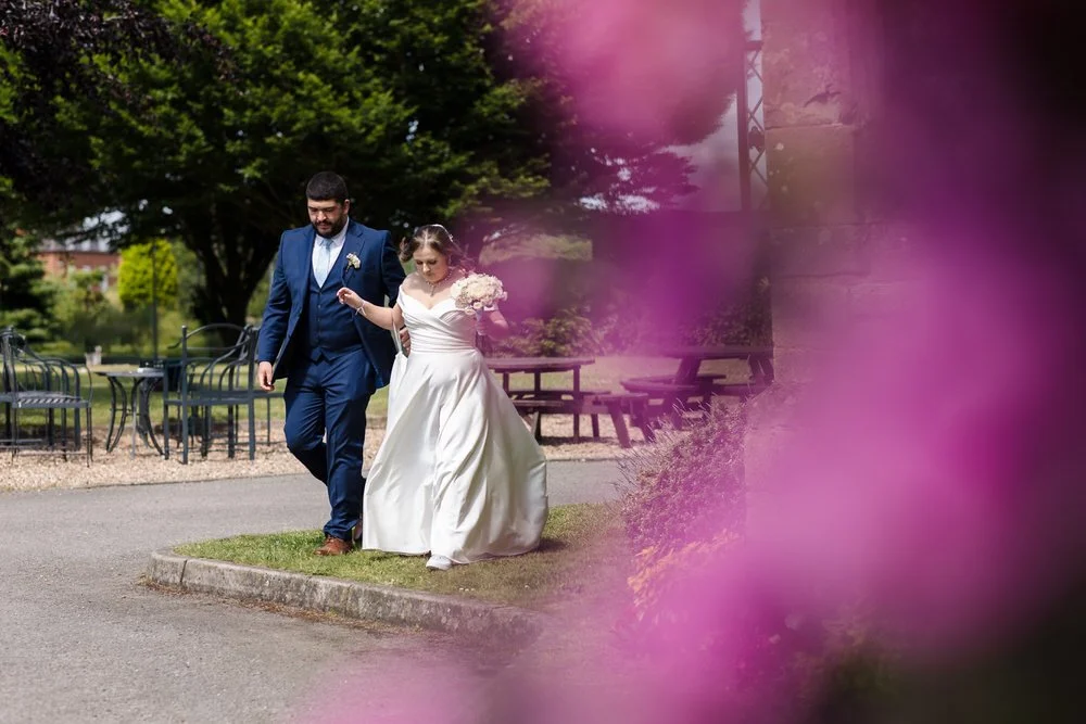 Candid walk through the gardens at Karma Salford Hall—bride holding bouquet, groom beside her, pink flowers in foreground.
