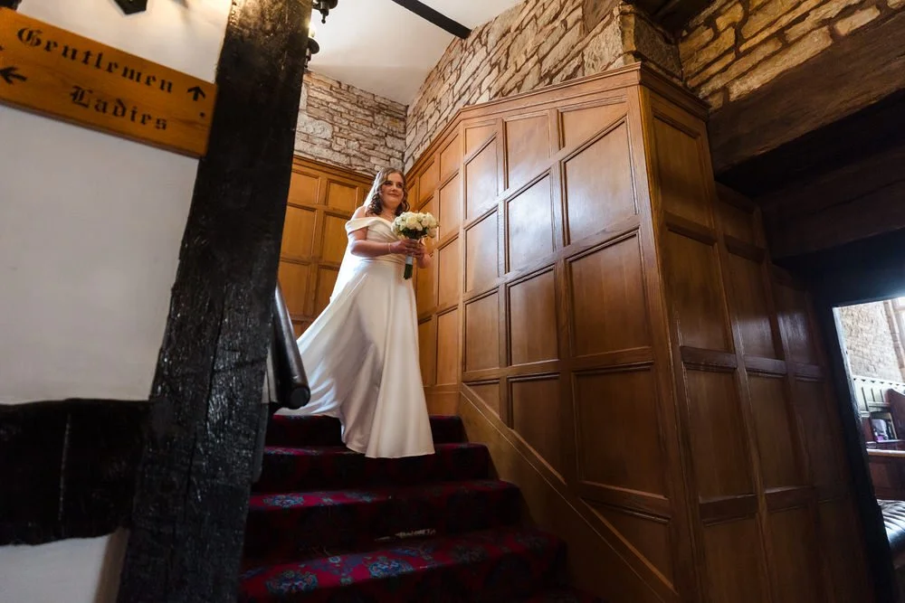 Bride descends the oak staircase holding her bouquet inside Karma Salford Hall before the ceremony.