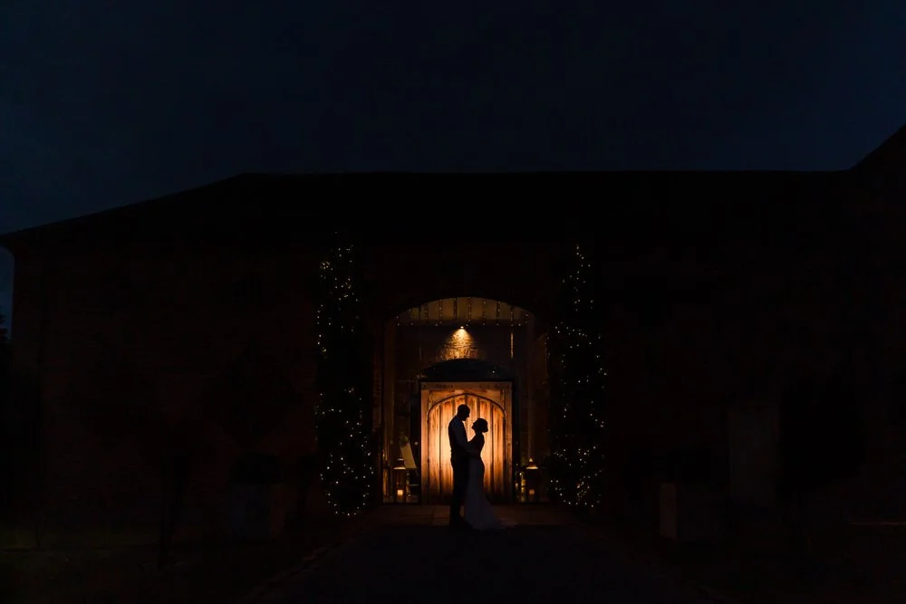 Dramatic night silhouette of bride and groom facing each other in the arched doorway of Shustoke Barn, glowing amber backlight and fairy-lit trees framing the couple against a deep blue evening sky