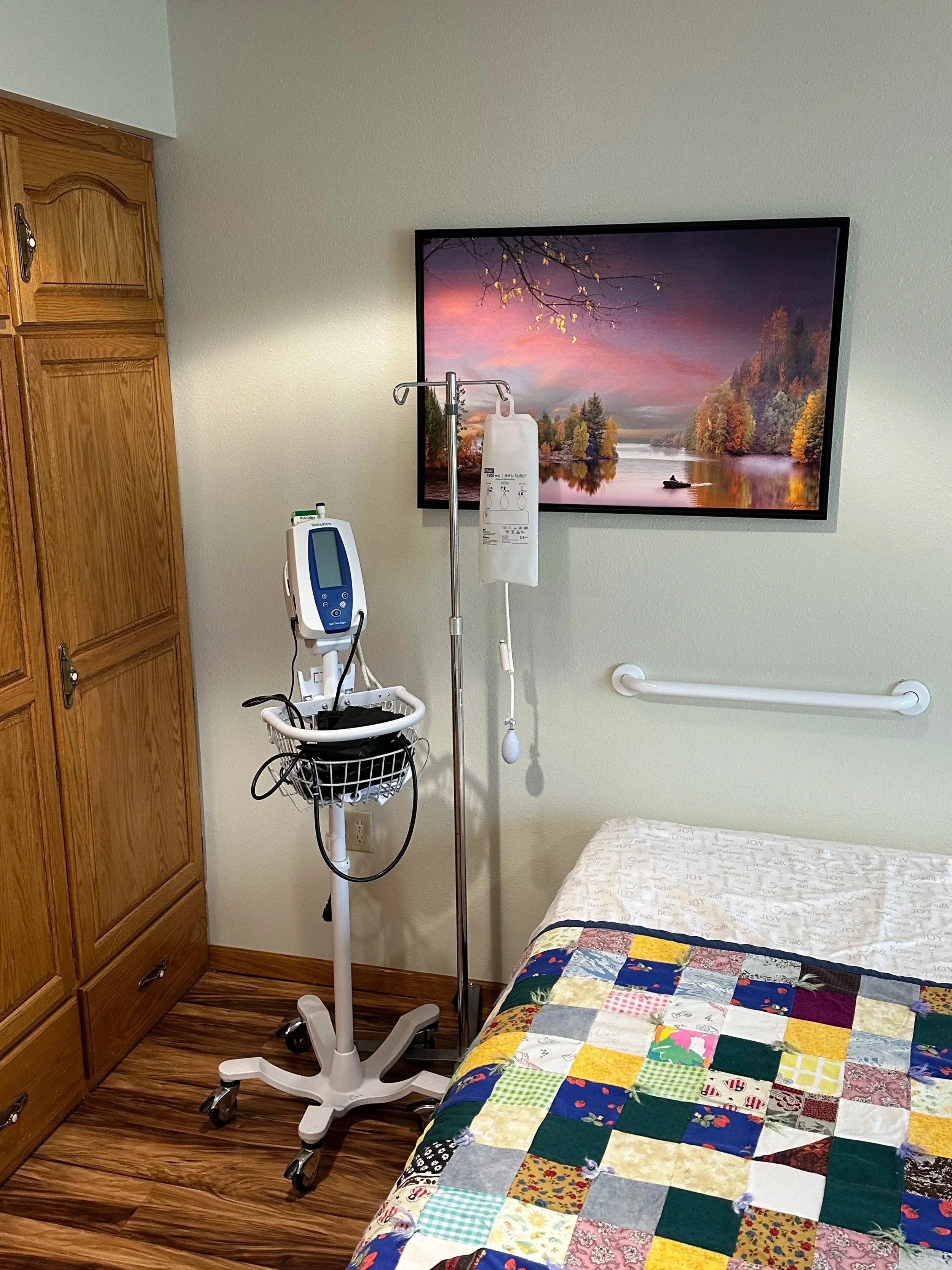 Hospital room with wooden storage cabinet, bed covered with a colorful patchwork quilt, wall-mounted television displaying a landscape scene with trees and water, medical monitors and IV pole next to the bed, wood-look flooring, and white walls.