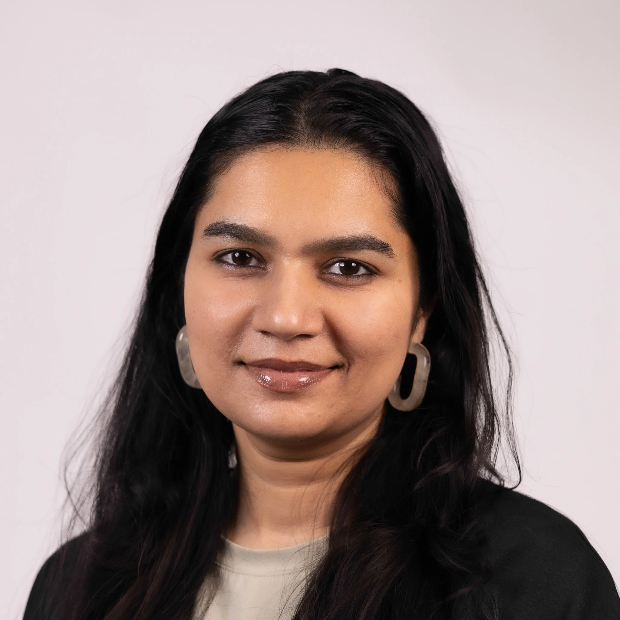 Portrait of a woman with long black hair, wearing pearl earrings and a black blazer, smiling softly against a white background.