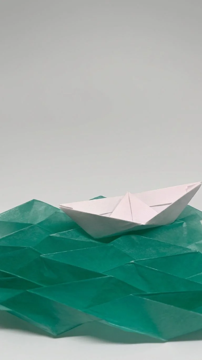 A small, white origami boat on green paper waves with a plain gray background.
