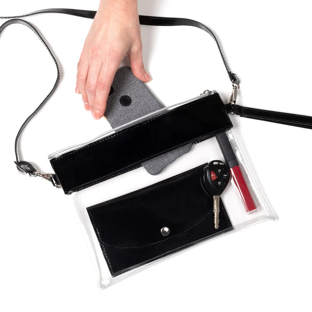 A clear purse with black trim seen from directly above. A hand reaches in from the upper left corner to remove a phone from the purse, which also contains a black matching wallet, red lip gloss, and car key.
