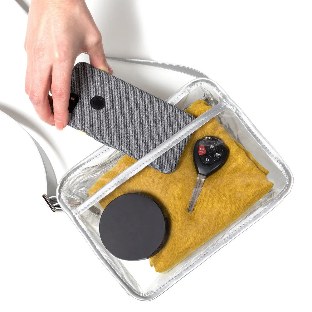 A clear purse with silver trim seen from directly above. A hand reaches in from the upper left corner to remove a phone from the purse, which also contains a black compact, yellow ochre scarf, and car key.