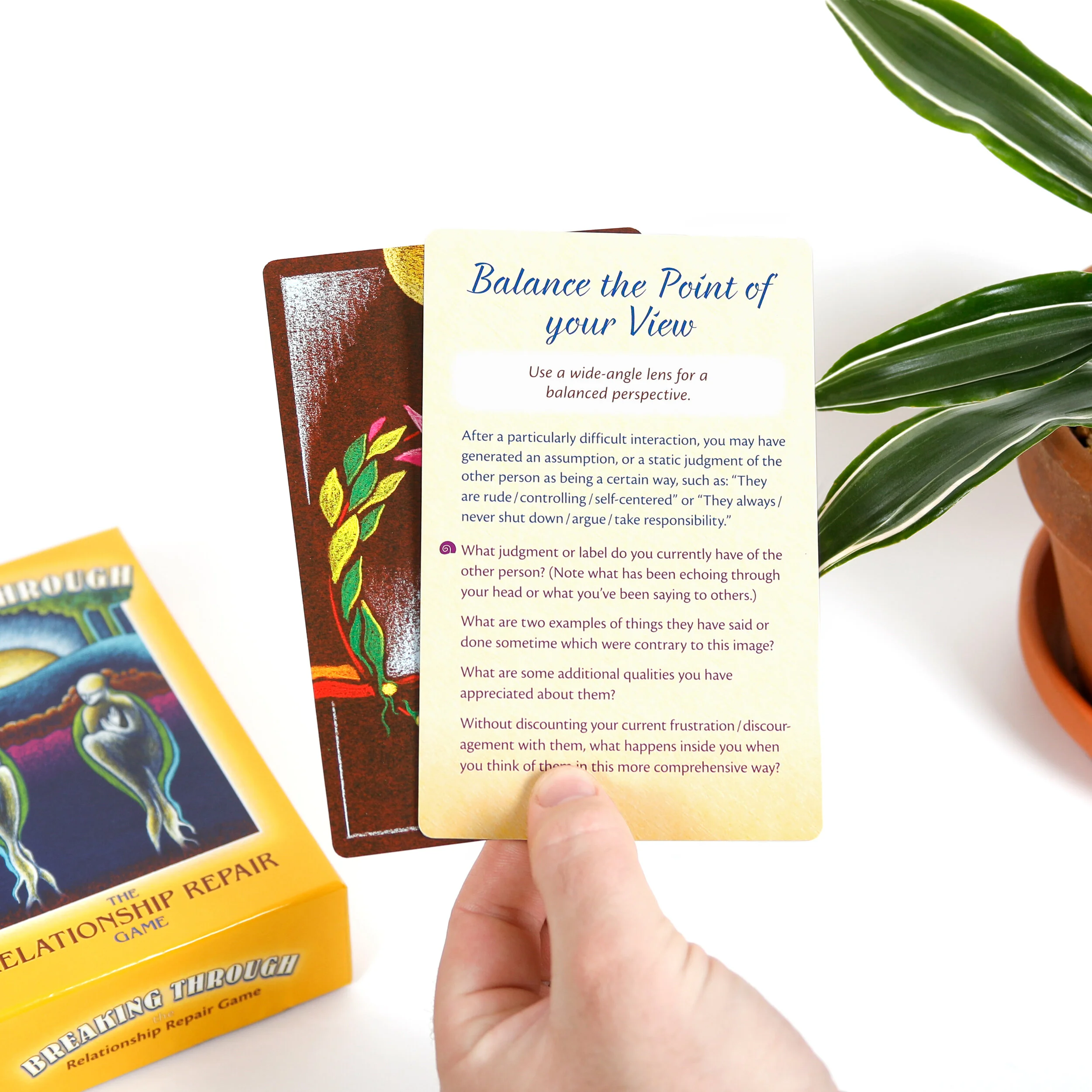 A relationship card game called Breaking Through, seen on a white background. A man's hand holds two cards, one showing artwork and one showing the back of the card with text. In the upper right of the image is a green plant in a terra cotta pot.