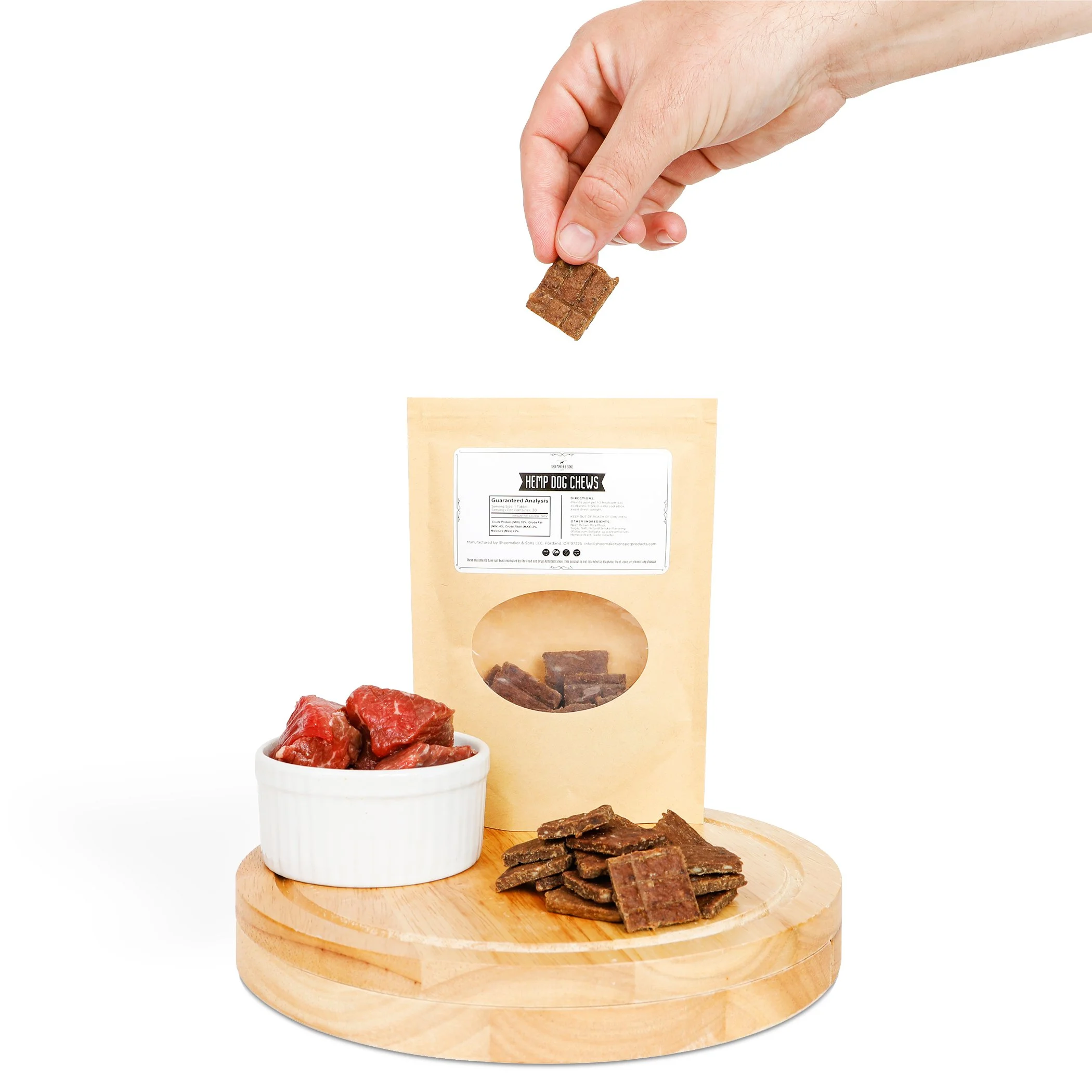 A tan package of CBD hemp dog treats sits on a light colored wood cutting board on white background. Treats can be seen through a plastic window on the front of the package and a man's hand removes a treat from the package from above. Next to the pac