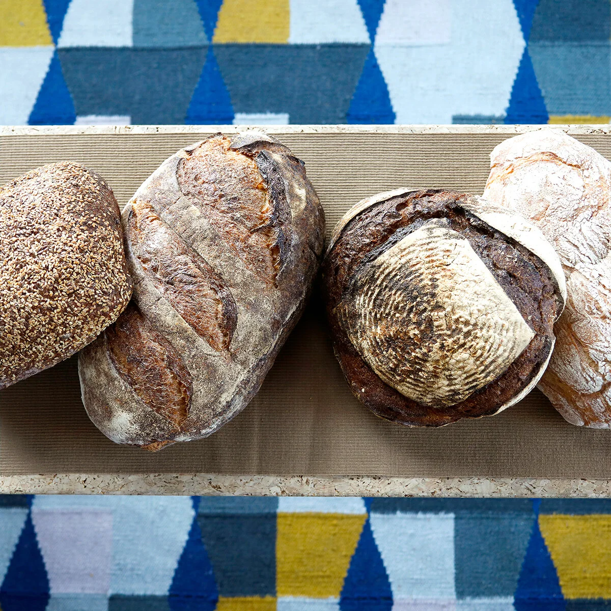 Rustic, artisan loaves of bread on a brown table runner seen from directly above. Below the table is a rug with blue and yellow geometric shapes.