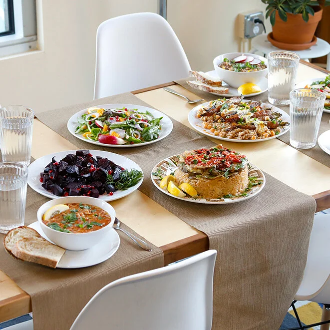 A table showing multiple vegan dishes. The food is colorful, with a beet dish, rice with tomatoes, salad, and lentils with lemon wedges on a table with brown runners and modern white chairs.