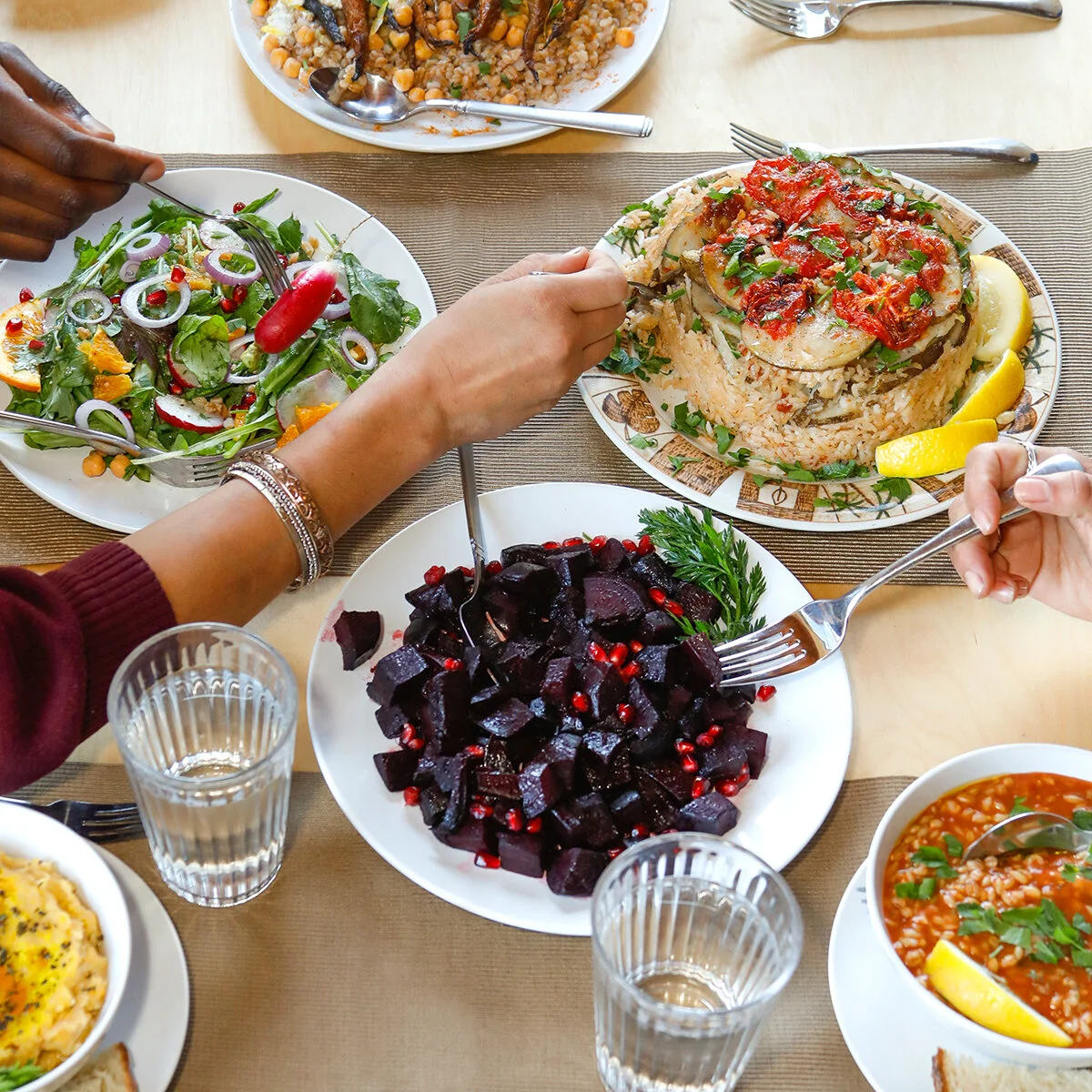 A table showing multiple vegan dishes with several hands reaching for helpings of food. The food is colorful, with a beet dish, tomatoes, and greens.