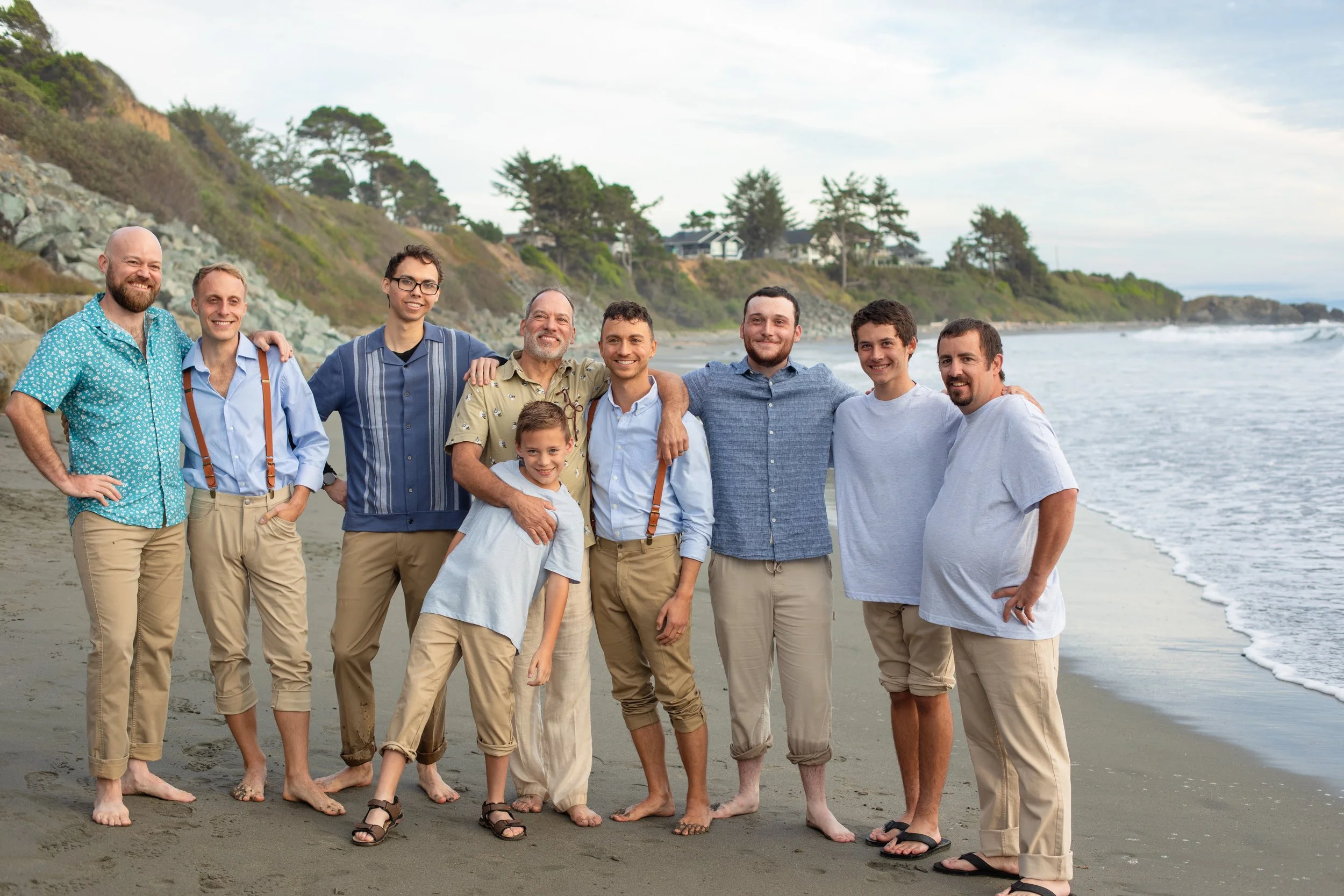 A group of nine men and one young boy standing shirtless with clothes, on a sandy beach near the ocean, with trees and houses on a hill in the background. Photo taken in Crescent City, CA.