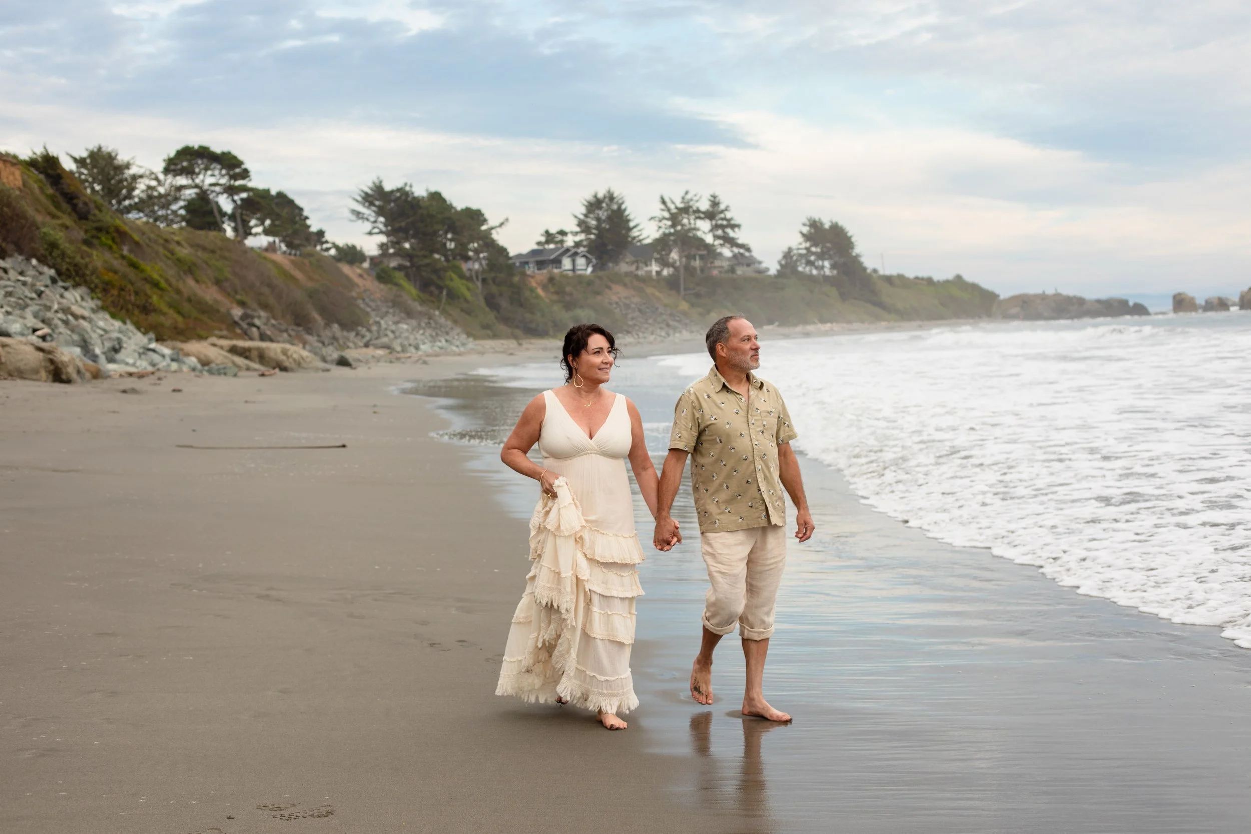 A couple walks barefoot along the shoreline of a beach in Crescent City, California holding hands, with waves gently crashing at their feet, overlooking cliffs with houses and trees under a partly cloudy sky.