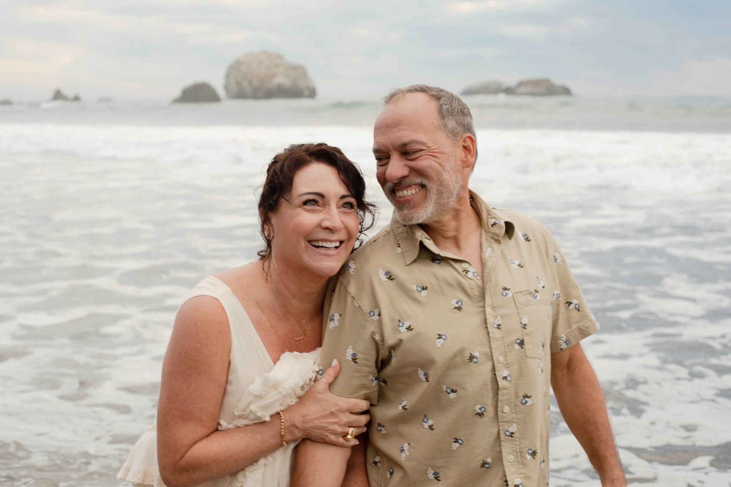 A smiling middle-aged woman and man enjoying time on the beach with ocean waves and large rocks in the background. Southern Oregon and Southern California Wedding Photographer.