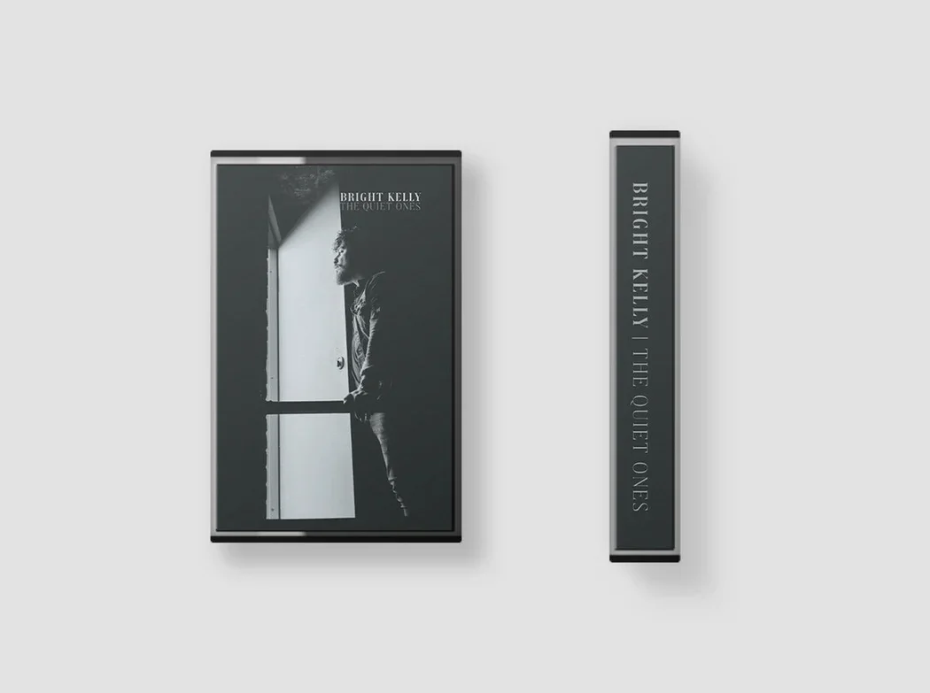 A cassette display for Bright Kelly's album 'The Quiet Ones'. The cover features a person standing by an open door, looking outside, in black and white. The case has the title and artist name in white text on a dark background.
