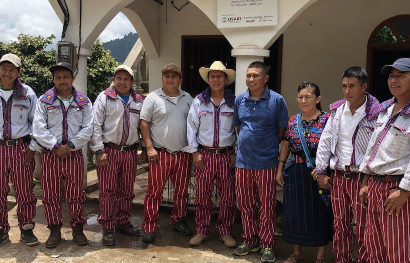 Group of nine people standing outdoors in front of a building. Seven men wearing traditional striped red and white pants, some with hats and jackets. Two women, one in traditional dress and the other in casual clothing. Mountain scenery in the background, cloudy sky.