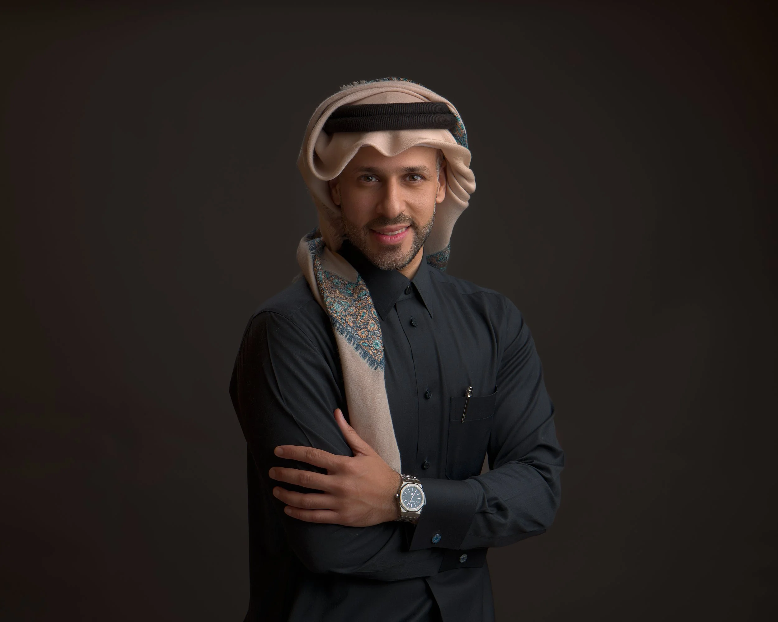A man wearing traditional Middle Eastern headgear, a black shirt, and a watch, smiling with arms crossed against a dark background.