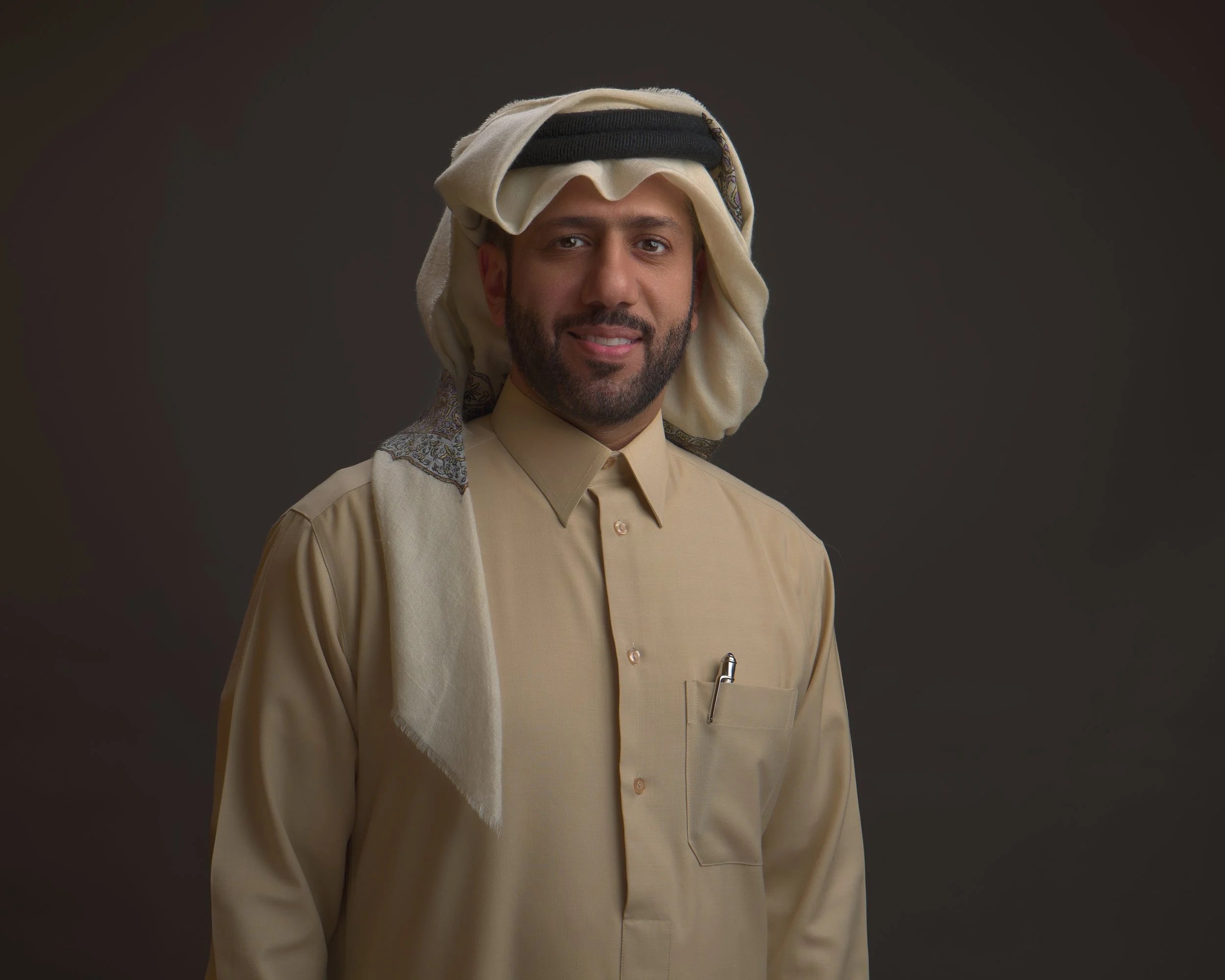A man with a beard wearing traditional Middle Eastern attire, including a beige thobe and a keffiyeh with an agal, smiling against a dark background.