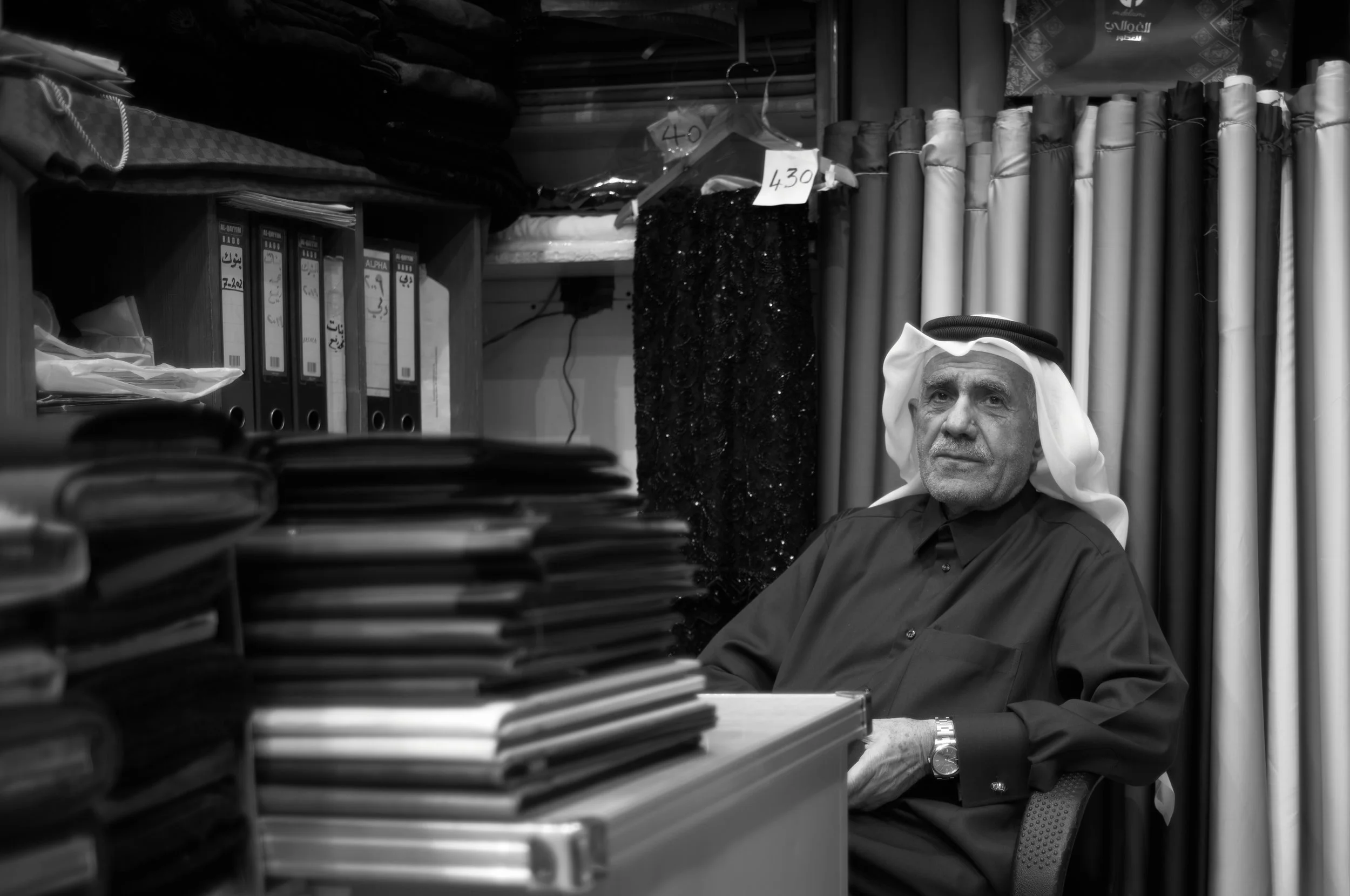 An elderly man wearing traditional Middle Eastern attire, including a head scarf, sitting behind a cluttered desk with numerous folders and documents, in a shop or office surrounded by fabric rolls and clothing items.