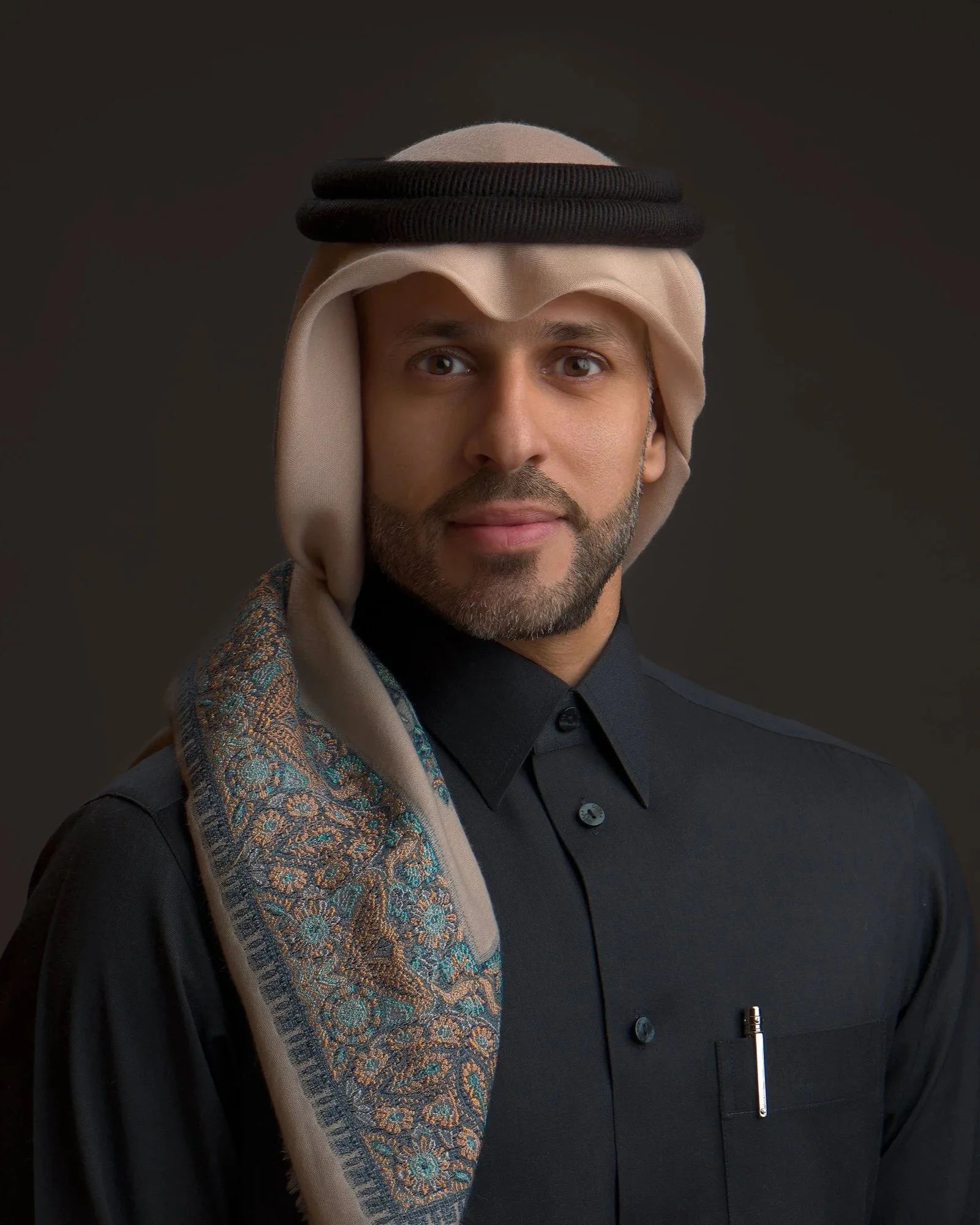 A man wearing traditional Middle Eastern attire, including a head covering with a black headband and a patterned scarf over his shoulder, dressed in a black shirt.