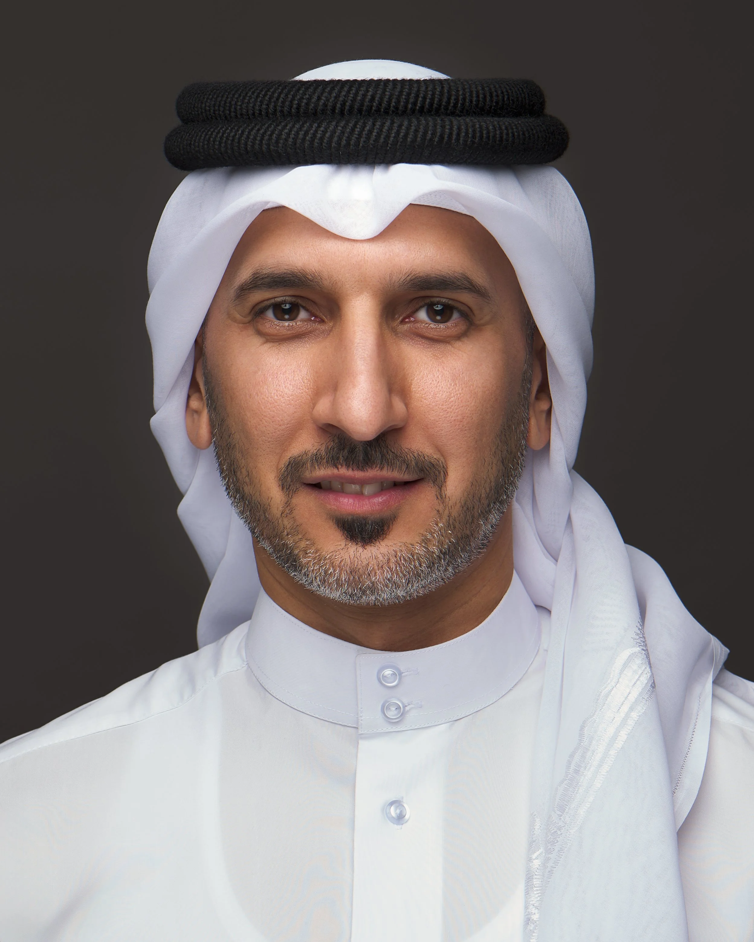 Close-up portrait of a Middle Eastern man wearing a white traditional thobe and headscarf with a black agal. He has a well-groomed beard and brown eyes, against a dark background.