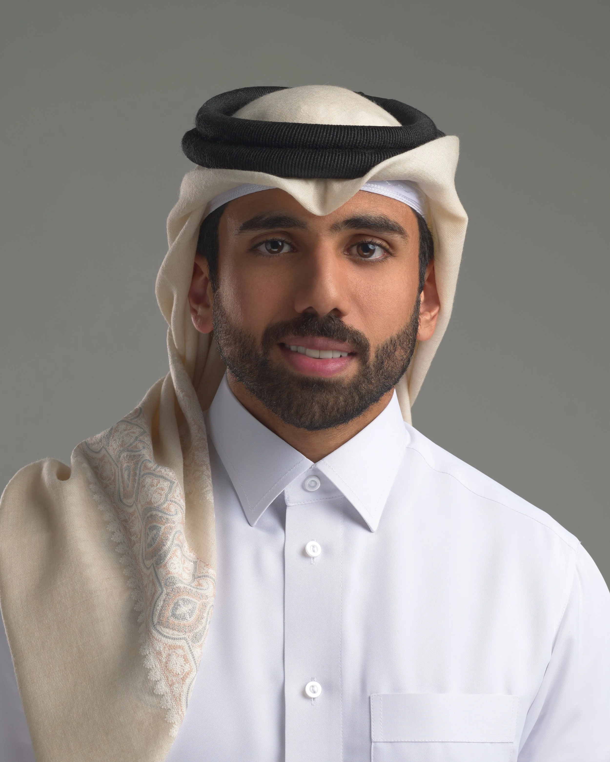 Portrait of a man wearing traditional Middle Eastern attire, including a white thobe, a keffiyeh with an agal, and a patterned shawl over his shoulder, against a plain gray background.