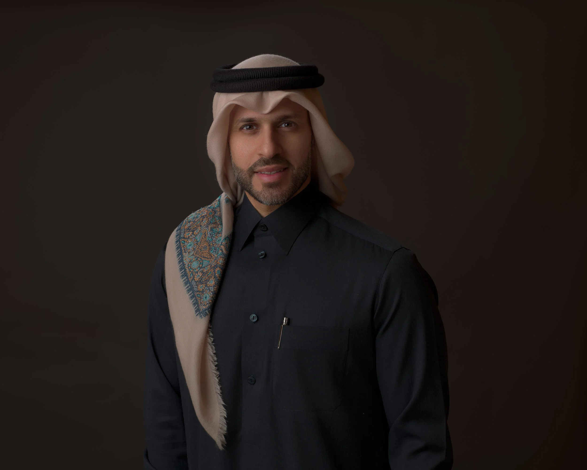 A man with a beard wearing traditional Middle Eastern attire, including a keffiyeh and a black thobe, standing against a dark background.