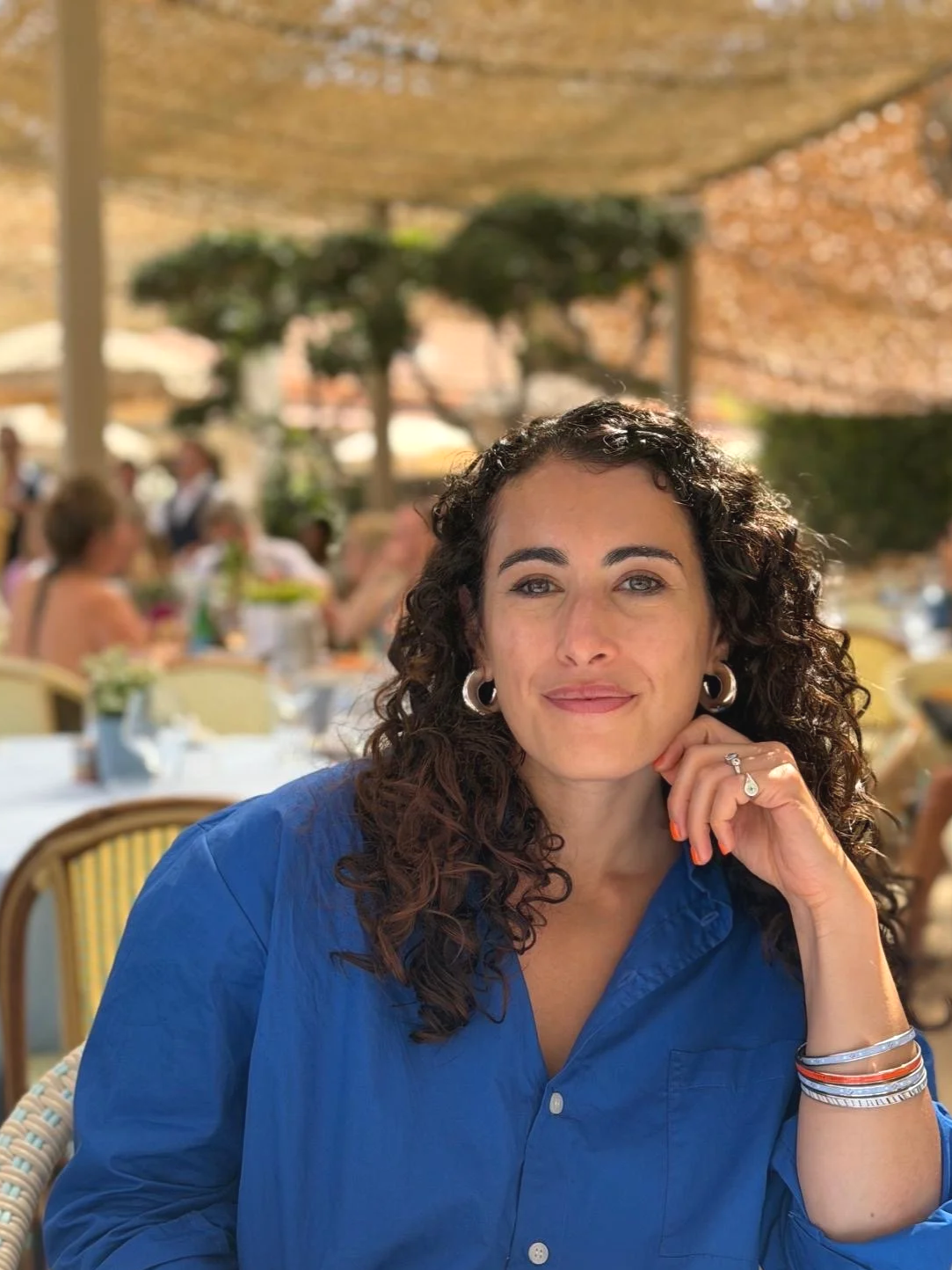 Lucy Morris, Strategy Director and Copywriter, pictured with dark, curly hair and earrings, wearing a blue shirt, sitting at an outdoor restaurant or cafe with other people in the background beneath a shaded canopy.