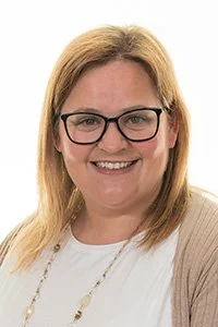 A woman with glasses and shoulder-length brown hair, smiling at the camera. She is wearing a beige cardigan over a black and white striped top.