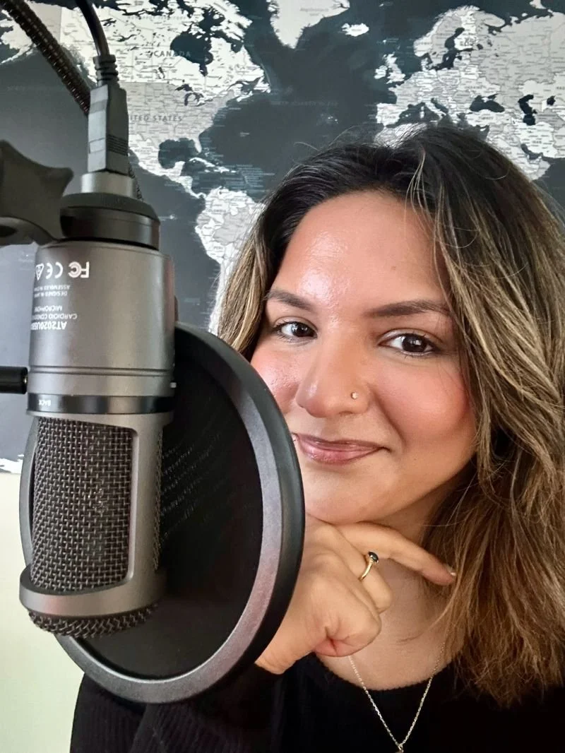 A woman with wavy brown hair and a nose piercing smiling and resting her chin on her hand, positioned in front of a large microphone with a pop filter, with a world map on the wall behind her.