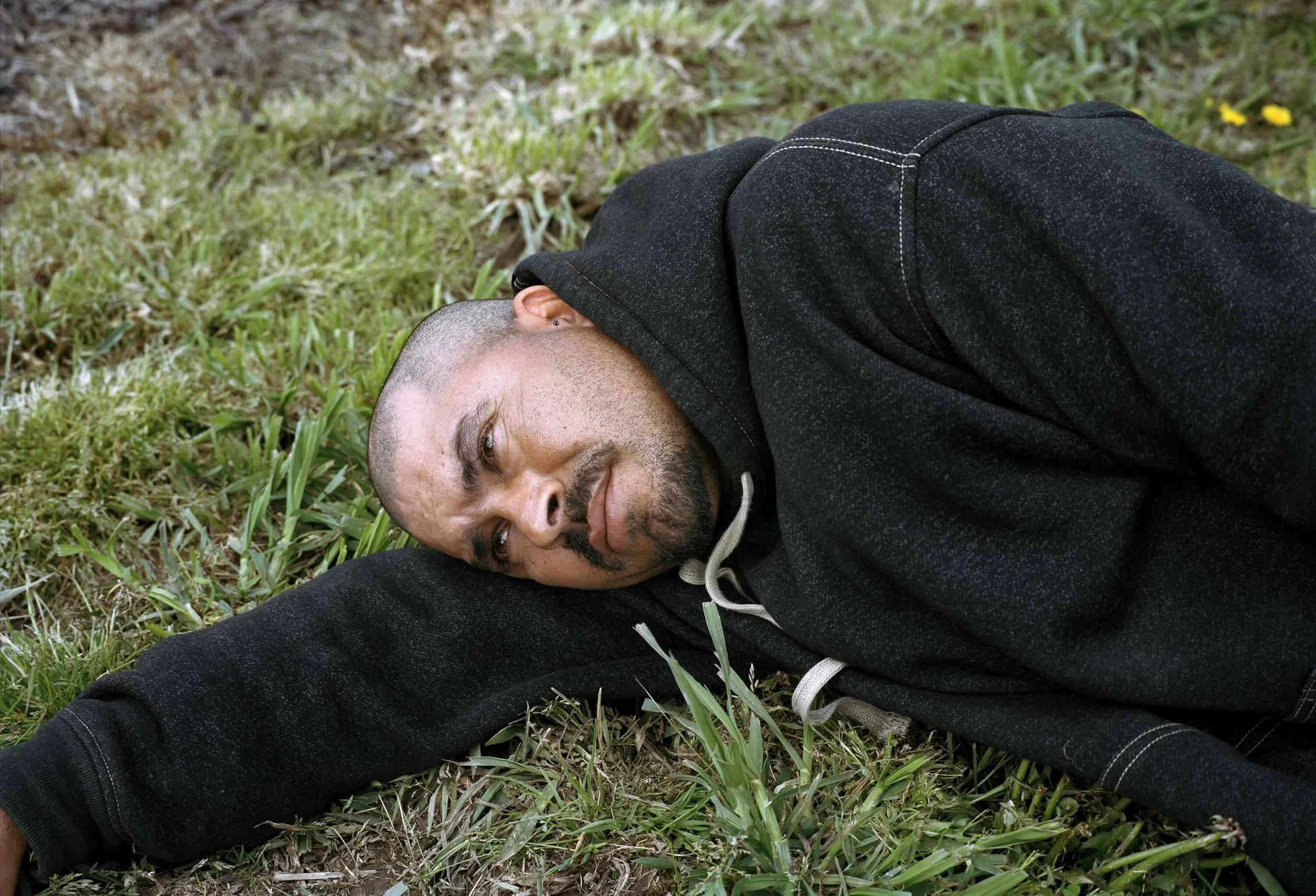A man in a black hoodie lays down on grass as he rests his head on his arm