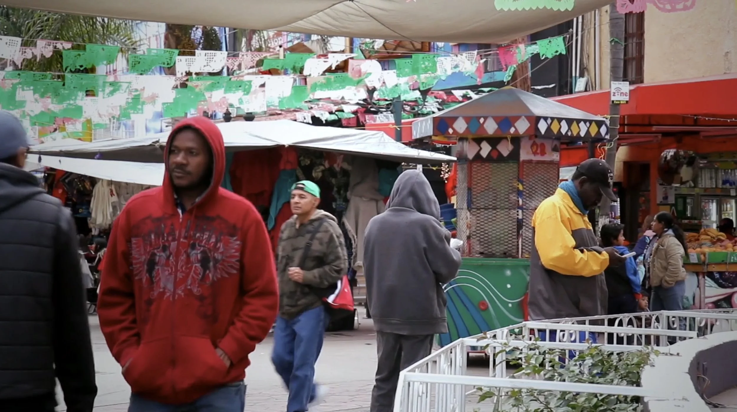 a film still of people walking through a plaza, with papel picado in the background