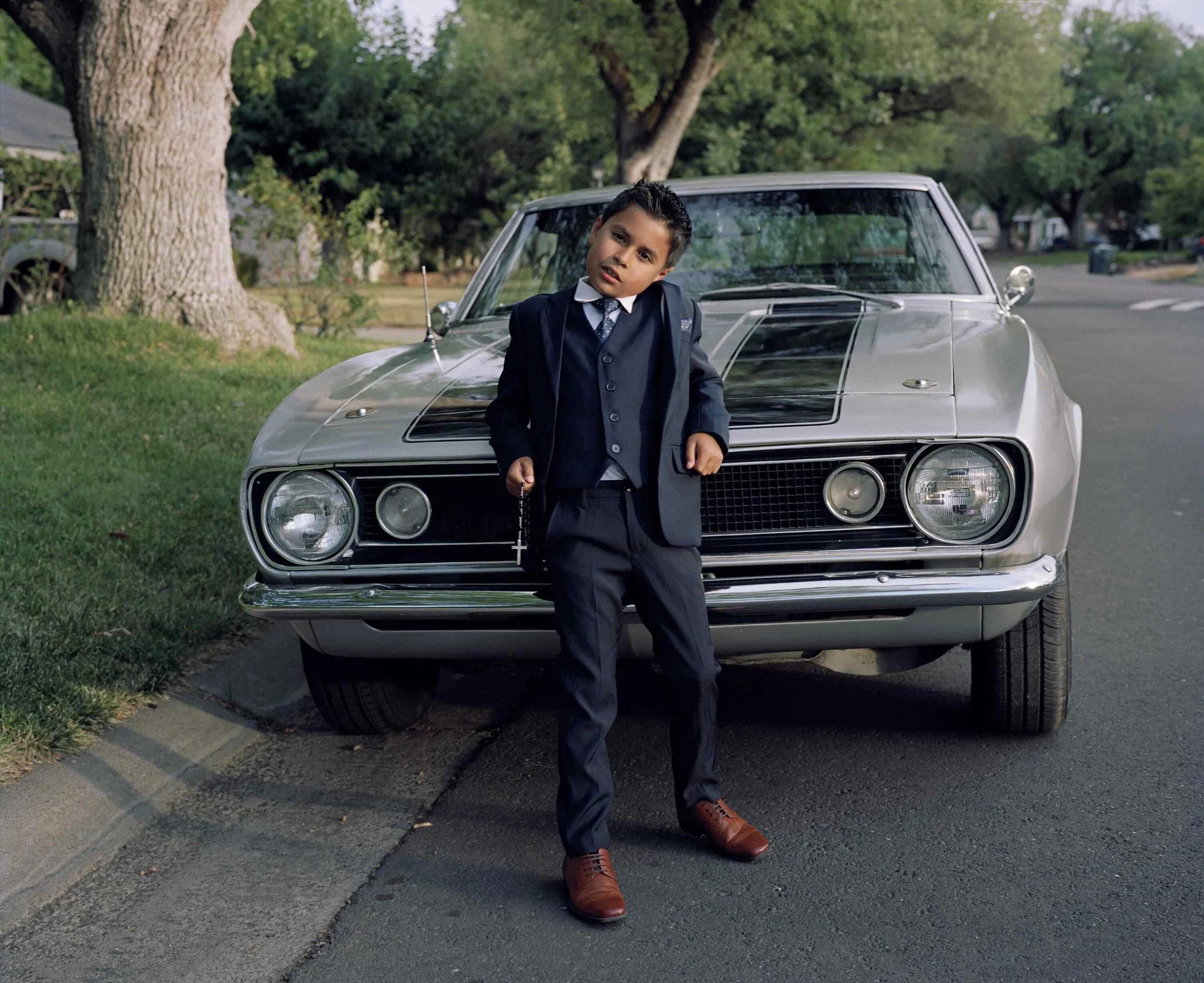 a young boy in a three-piece suit poses in front of a camaro