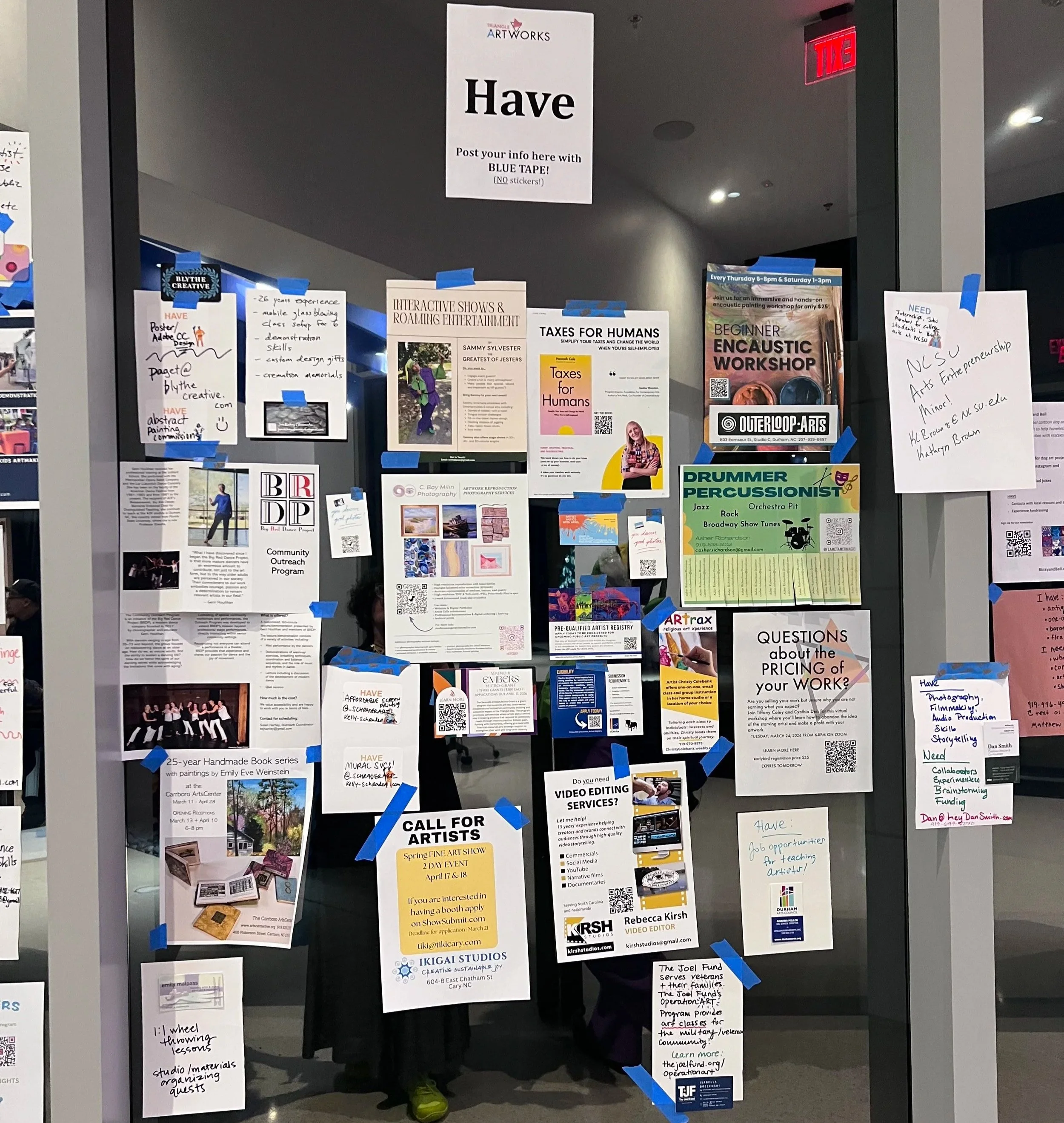 An image of a glass wall with many pieces of paper, printed with requests or offers for studio space, support, jobs, gigs, work and grants for artists and performers taken at SMASH! 2026 in North Carolina