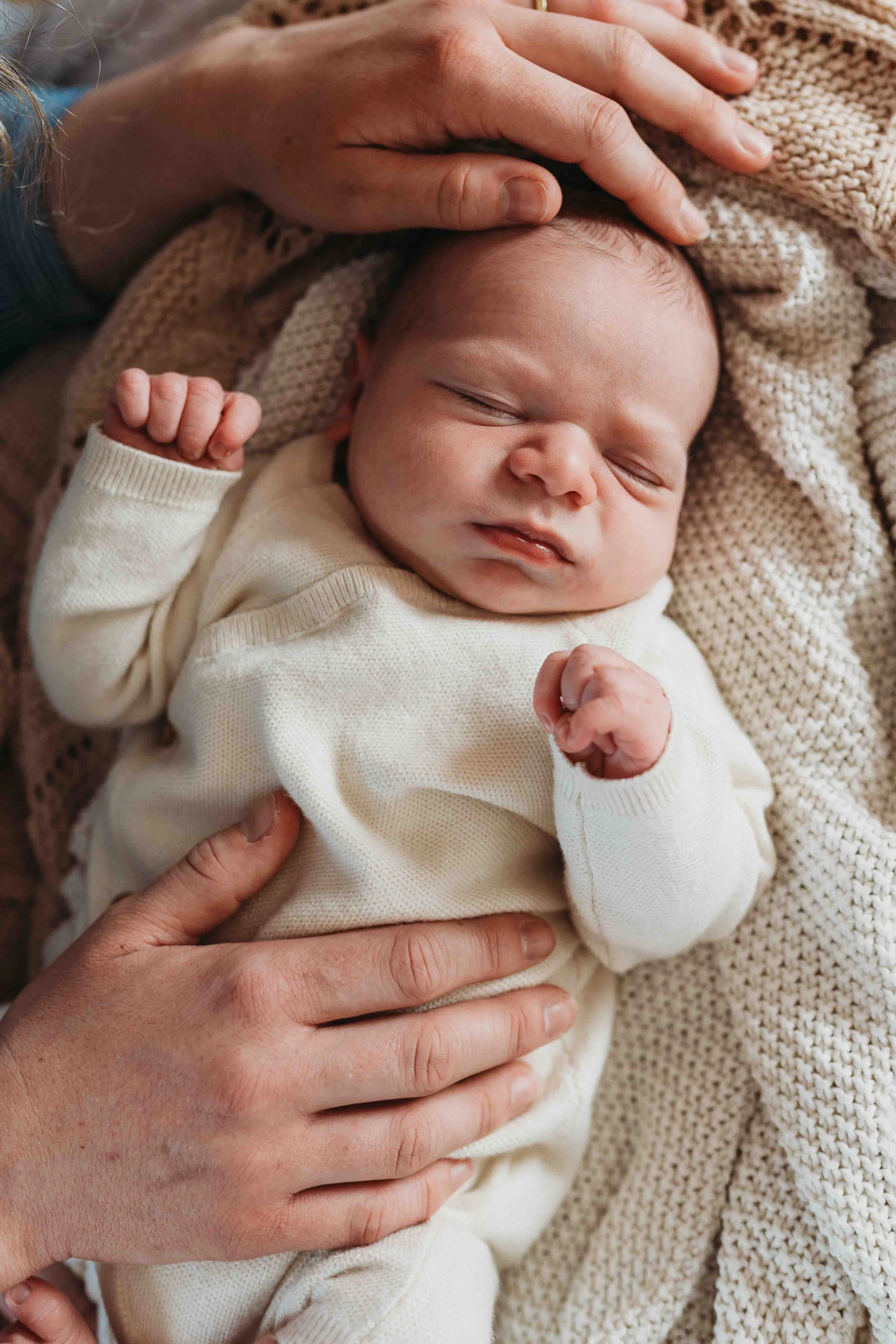 newborn baby resting on neutral knit blankets with a parent’s hand gently touching their head
