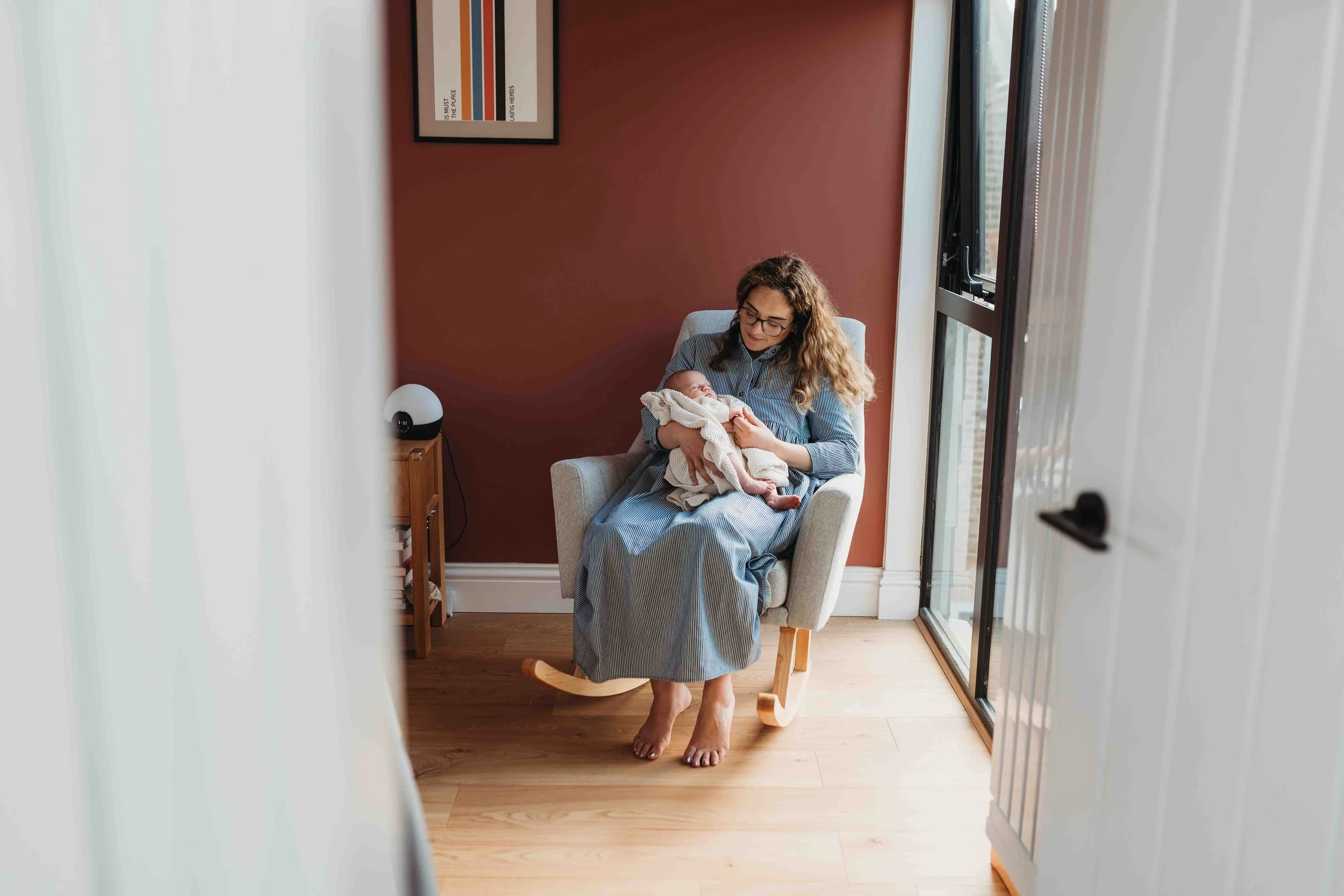 mother holding newborn baby while sitting on a rocking chair in a light-filled nursery during an at-home newborn photoshoot