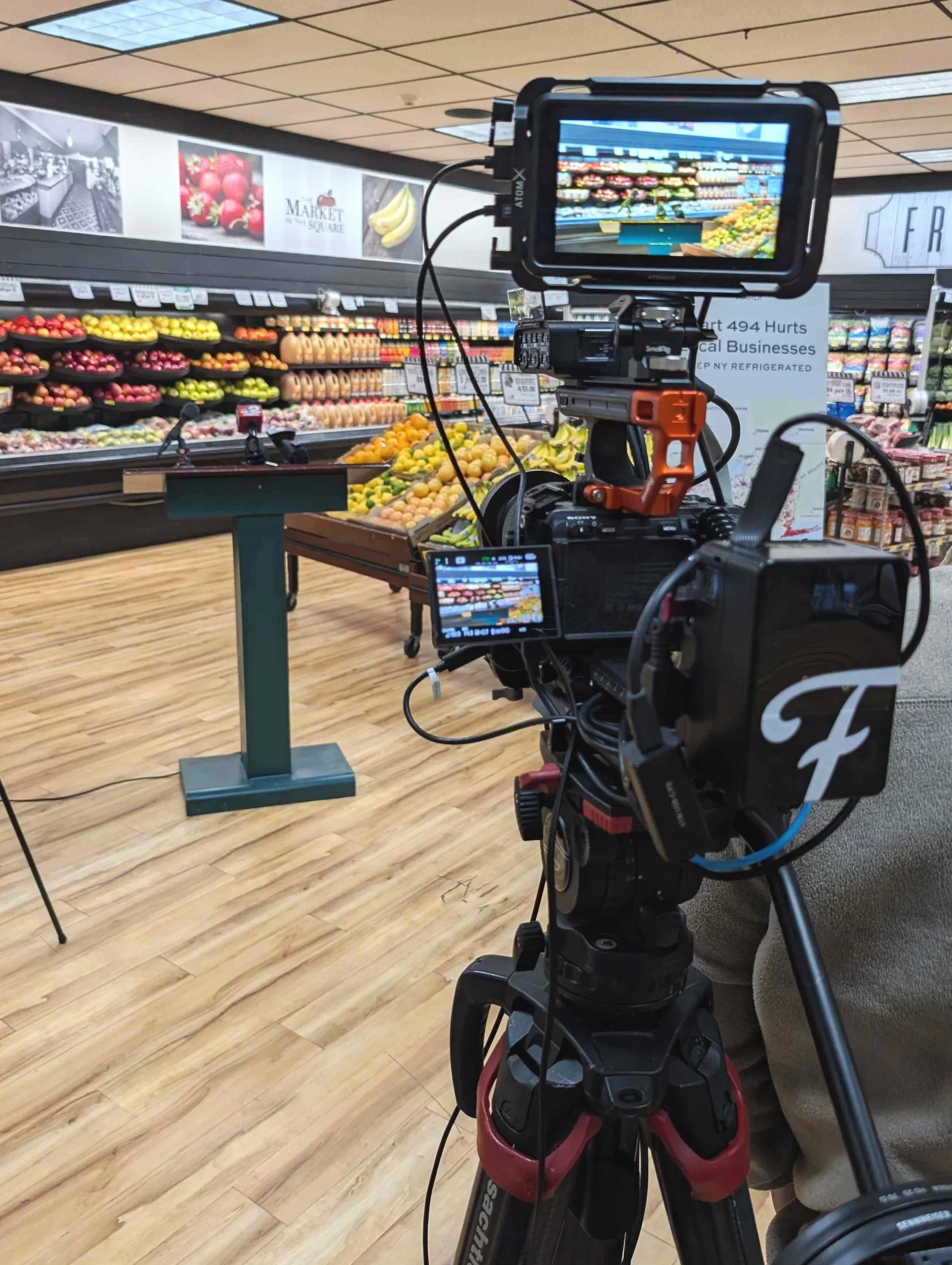 Professional Camera on Tripod, setup for Live Streaming of political speaking session, background is a produce section at a grocery store