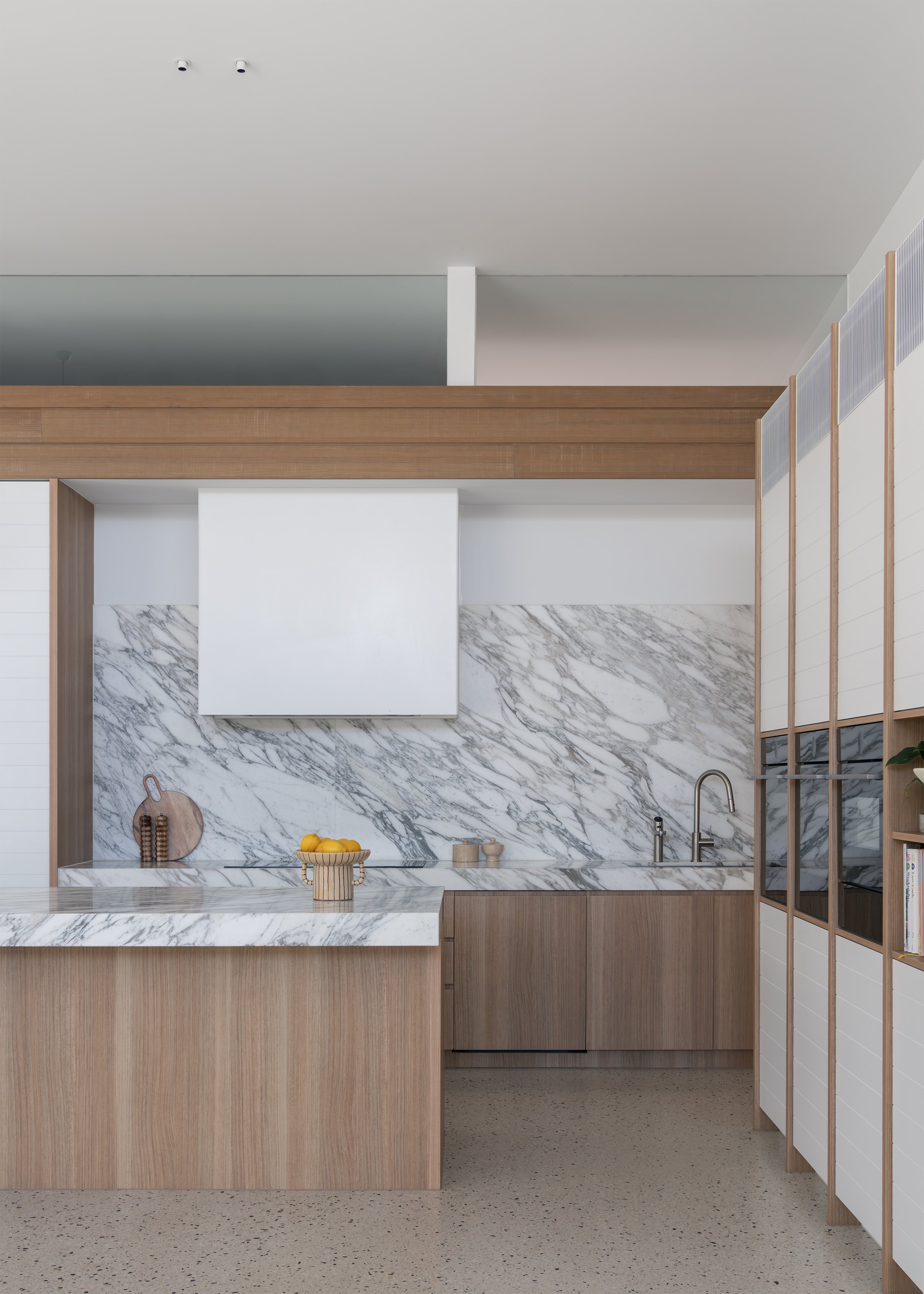 Kitchen at Channel framed by timber cabinetry and veined stone surfaces, opening toward a quiet concrete-lined passage.