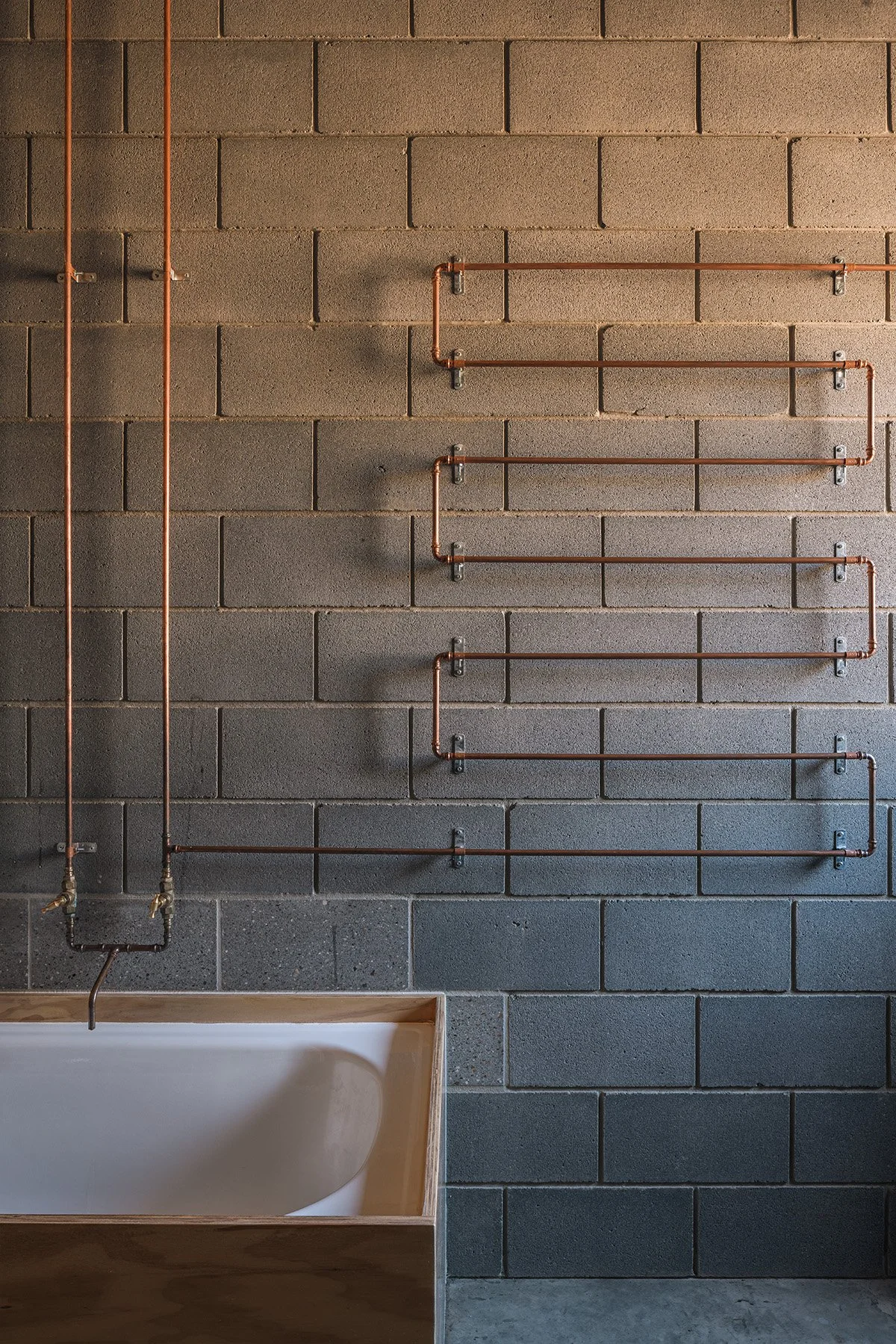 Exposed copper plumbing arranged across a concrete block bathroom wall above a built-in bath at Mansfield House.