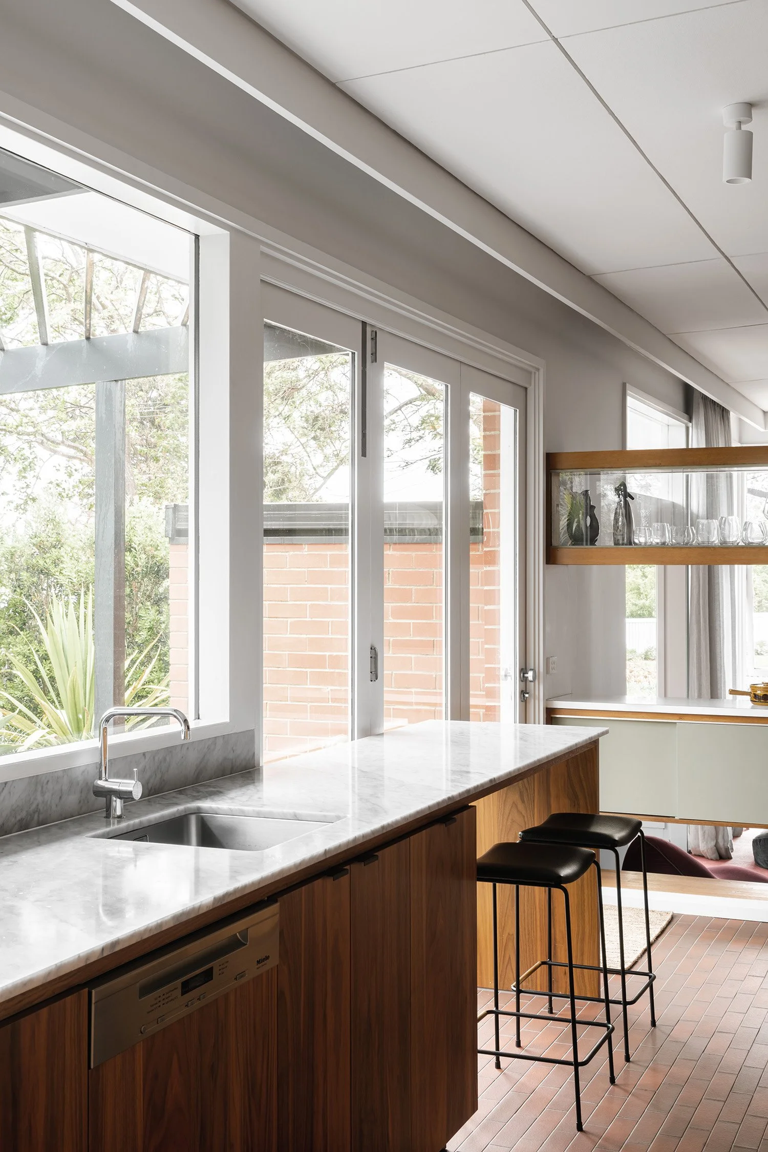 Mid century modern breakfast bar with black stools, a pale benchtop and a glass display cabinet beside garden-facing windows.