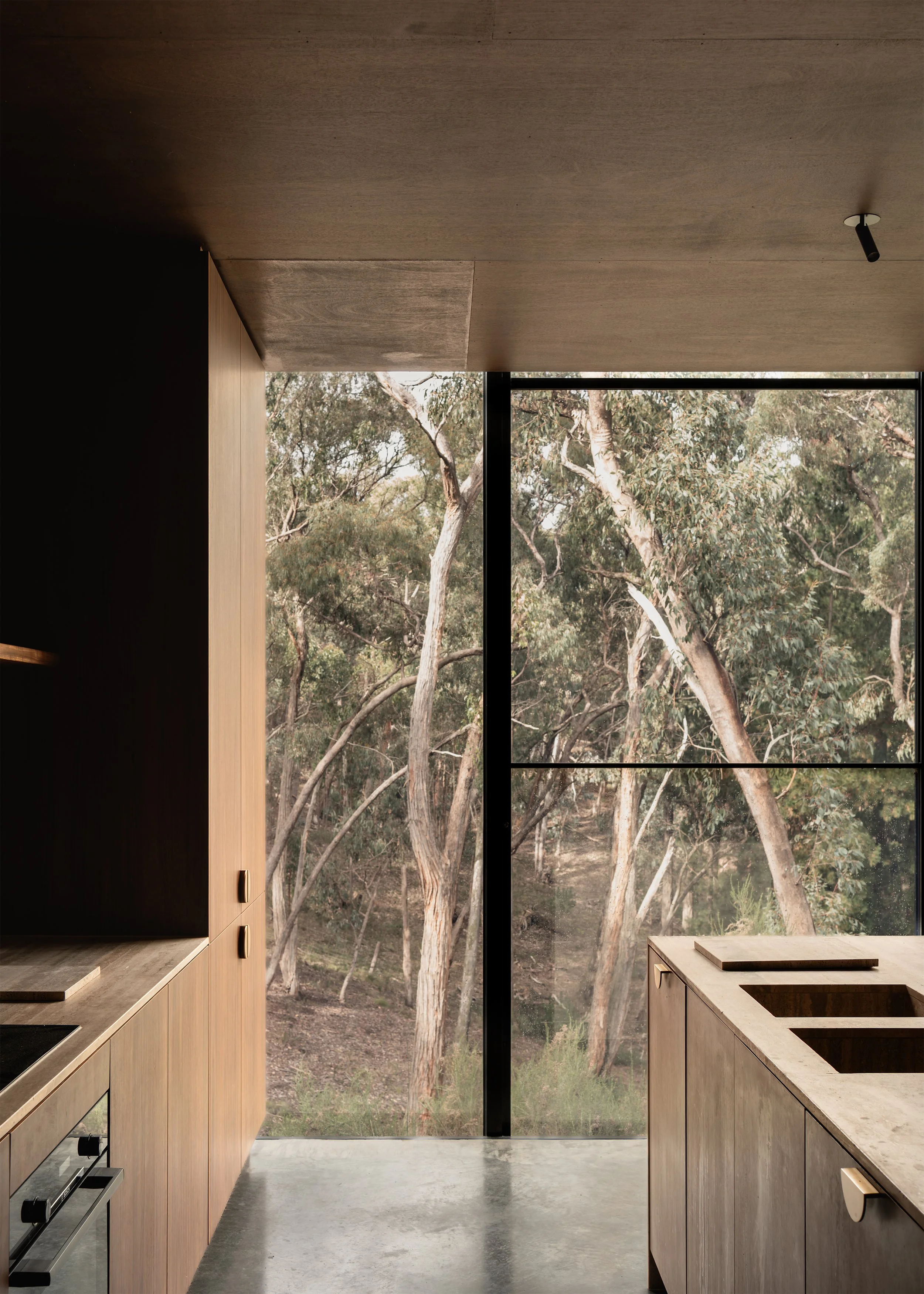 Kitchen at Matchbox framed by timber joinery, travertine stone benchtops and a full-height window to the eucalypt landscape.