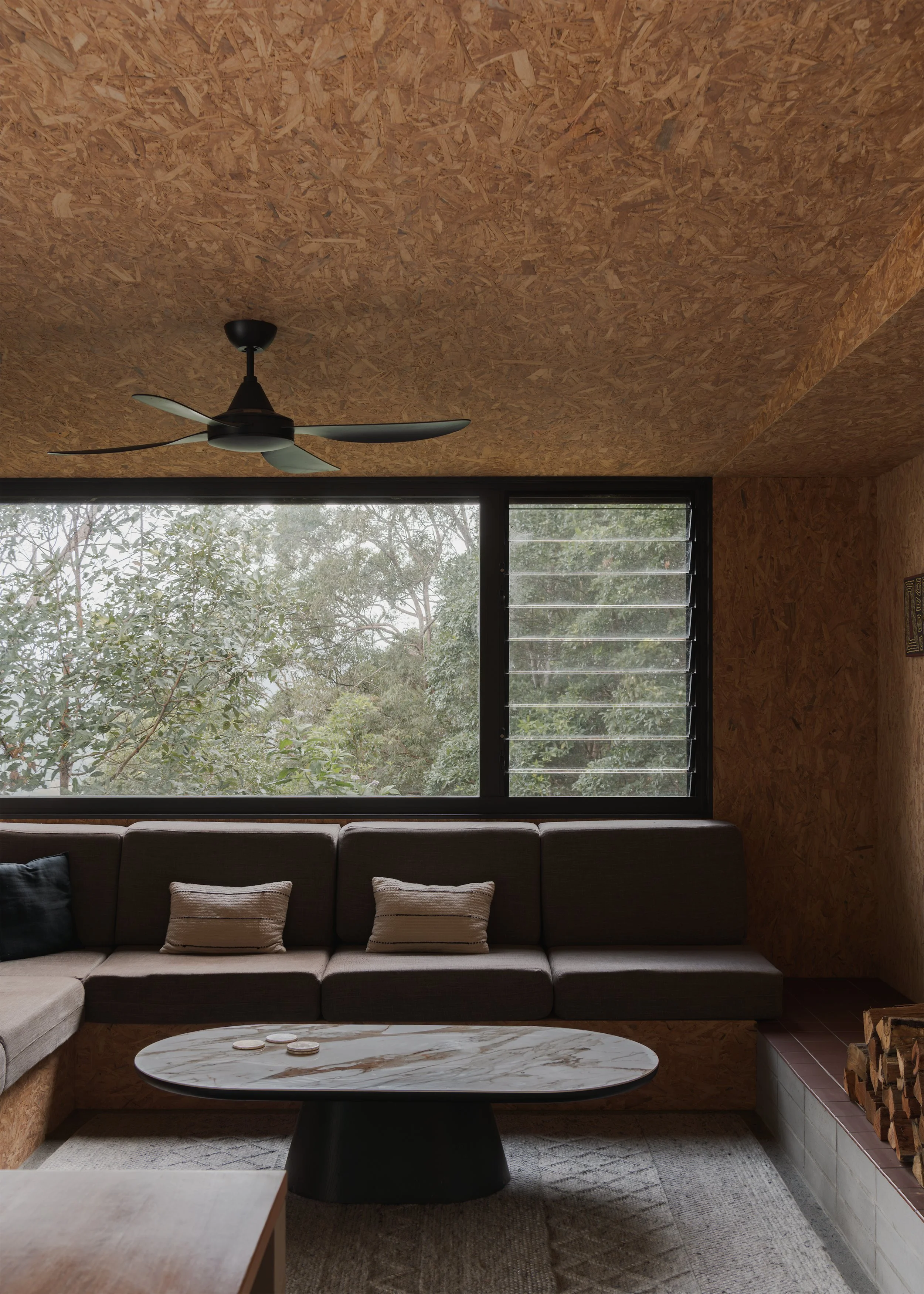 Built-in lounge at Tallowwood Cabin beneath a broad window to the eucalypt canopy, with exposed OSB and a low stone coffee table.