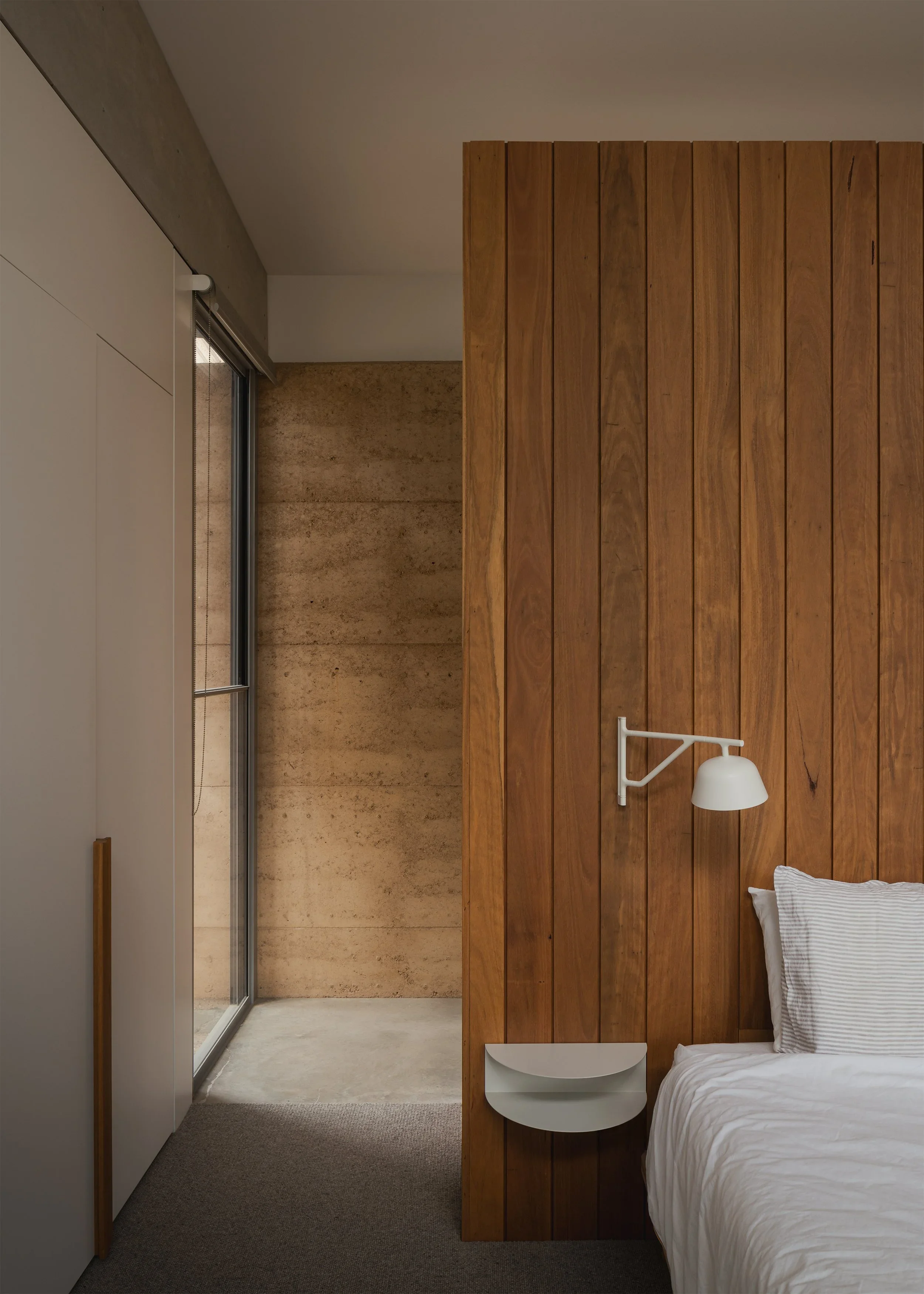 Timber-panelled bedhead beside a rammed-earth wall at Forest Road House, with a white reading light, floating shelf and narrow window.