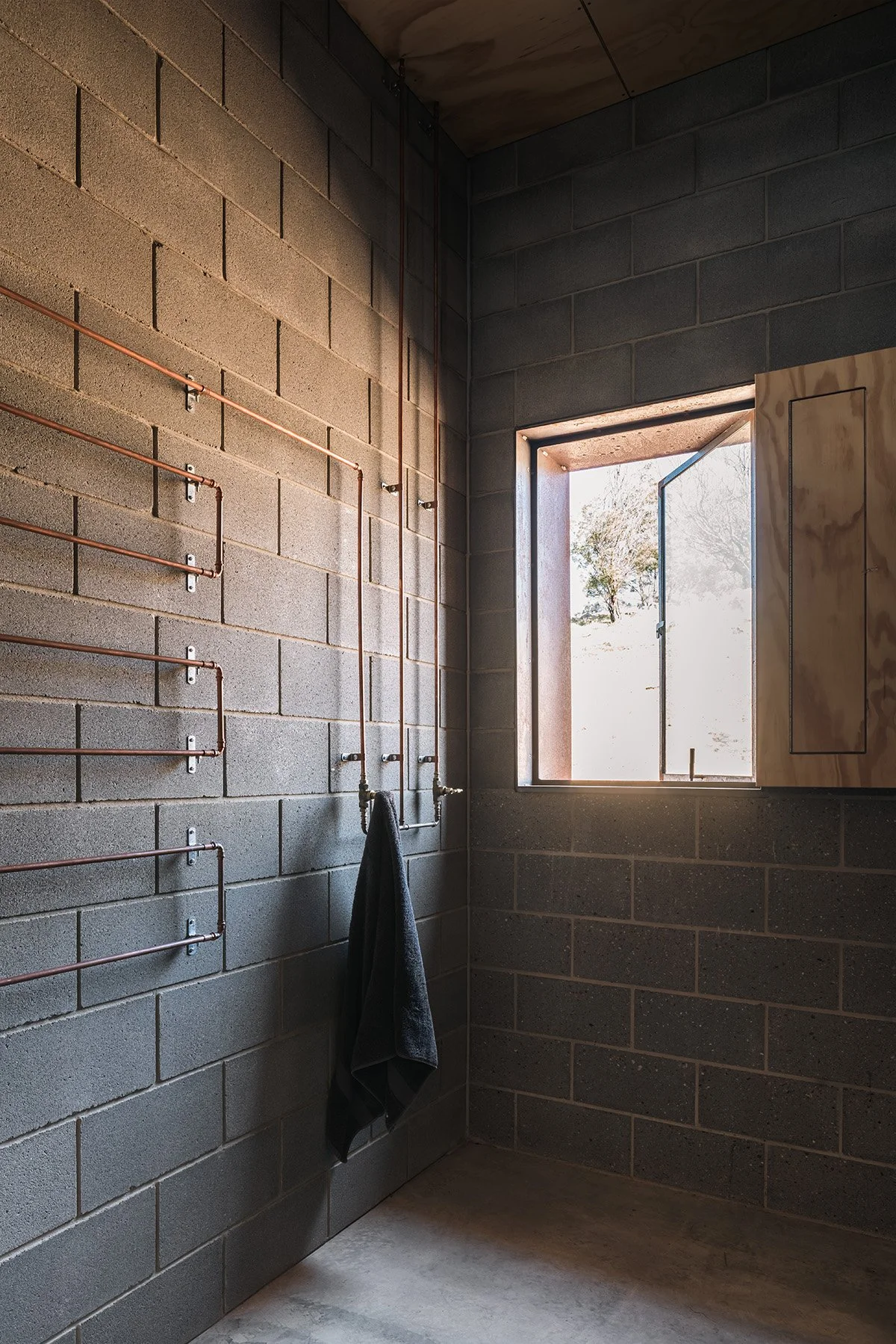 Concrete block bathroom at Mansfield House with exposed copper pipework, plywood joinery and an open window to the rocky site.