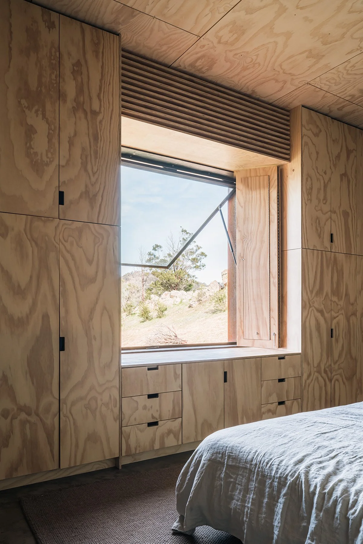 Minimalist bedroom lined with plywood wardrobes and a built-in window seat overlooking the rocky Mansfield hillside.