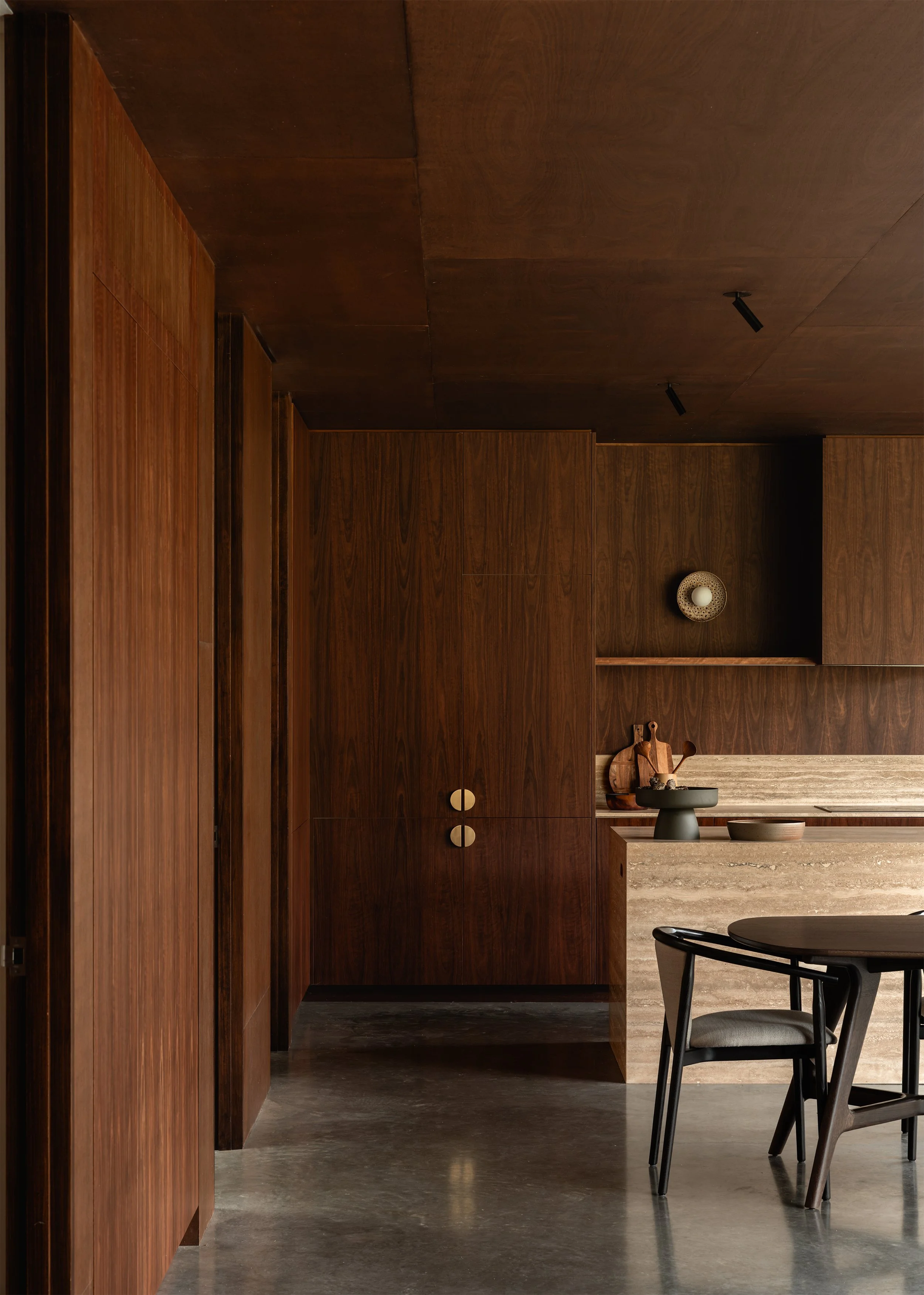 Dark timber joinery lining the kitchen and circulation space beside the travertine stone island and dining area.