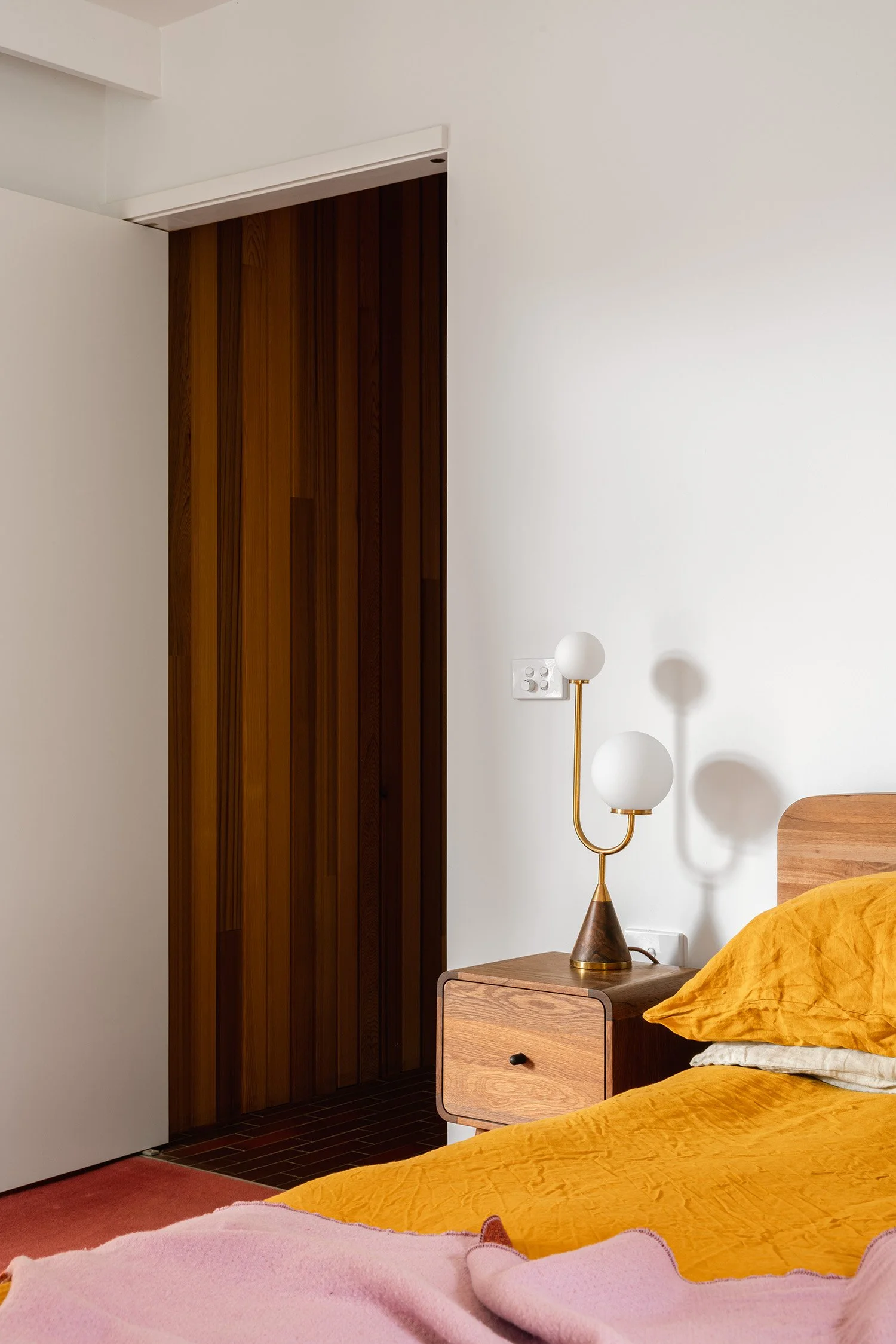 Mid century modern bedroom detail with mustard bedding, a timber bedside cabinet and a brass lamp with two white globes.