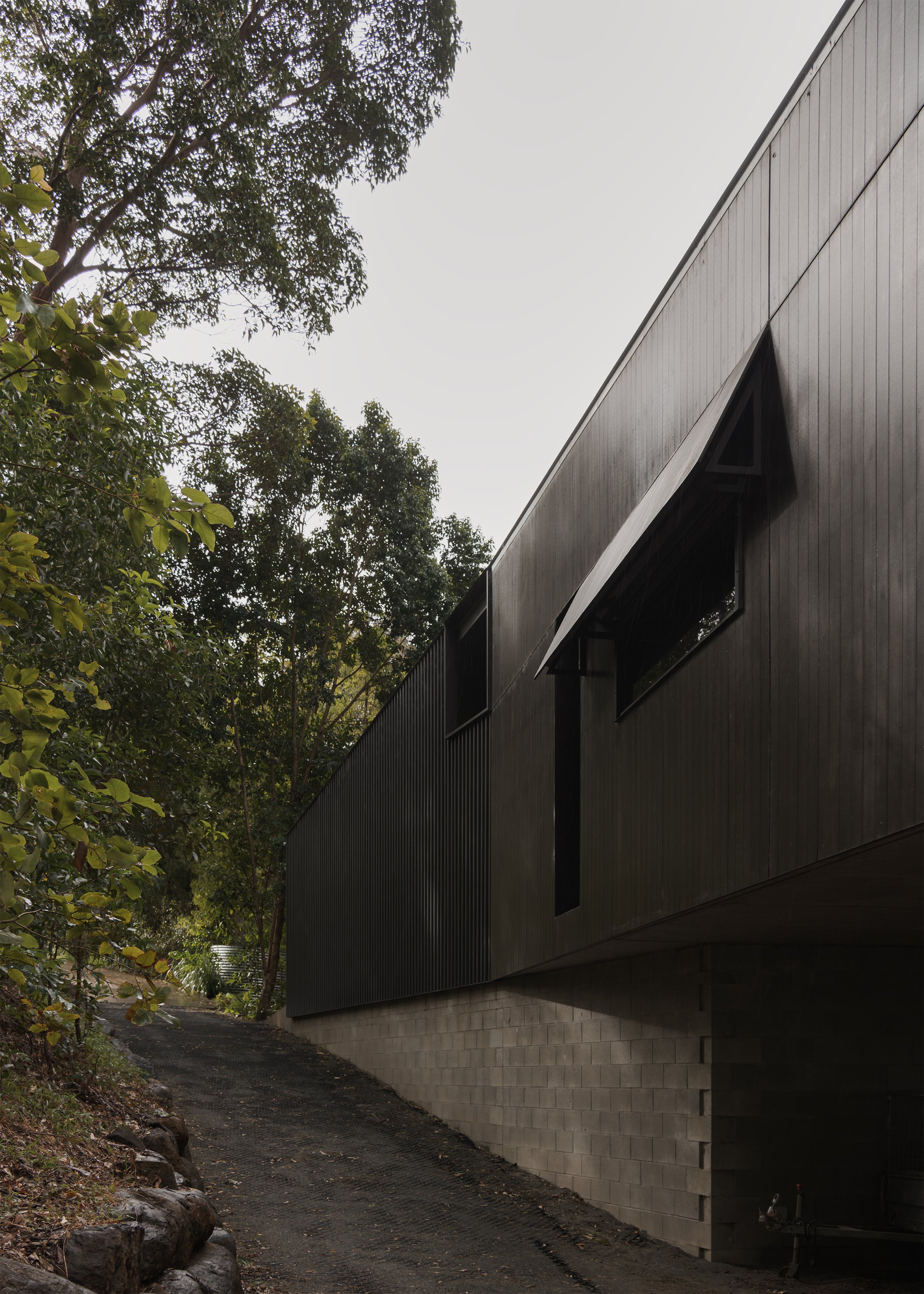 Elevated side elevation of Tallowwood Cabin showing dark INEX>Express cladding, automatic bushfire shutters and a concrete-block undercroft.