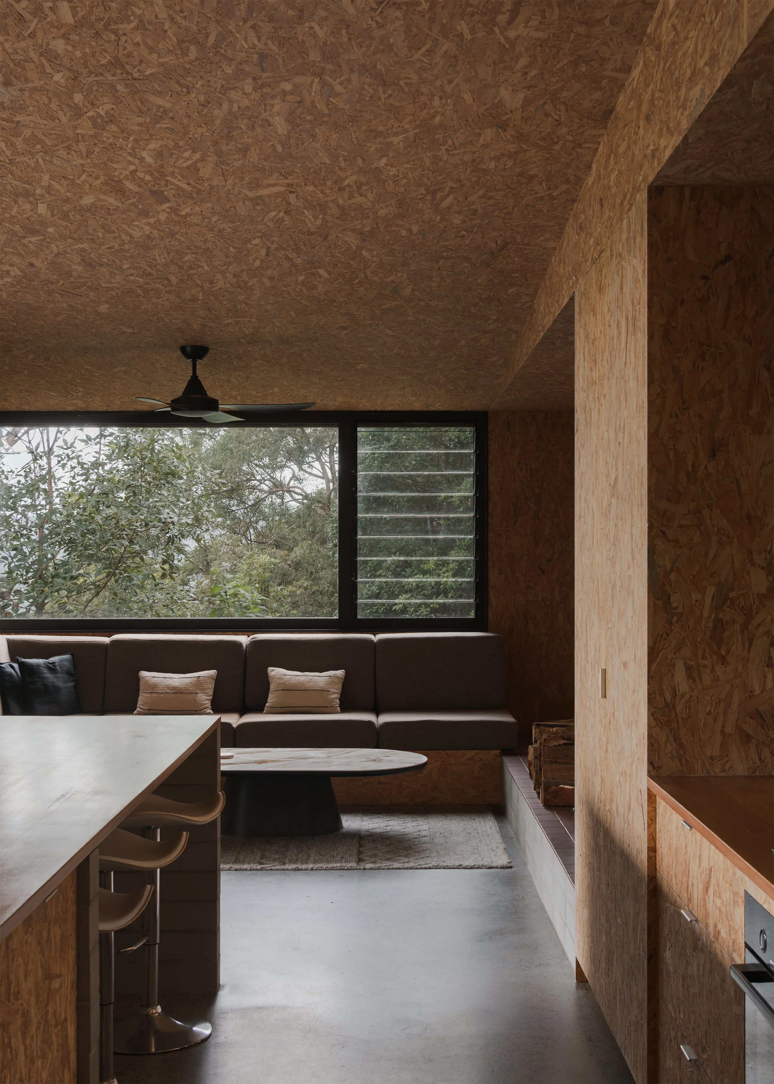 Compact kitchen and living area at Tallowwood Cabin, where exposed OSB, built-in seating and polished concrete create a restrained interior.